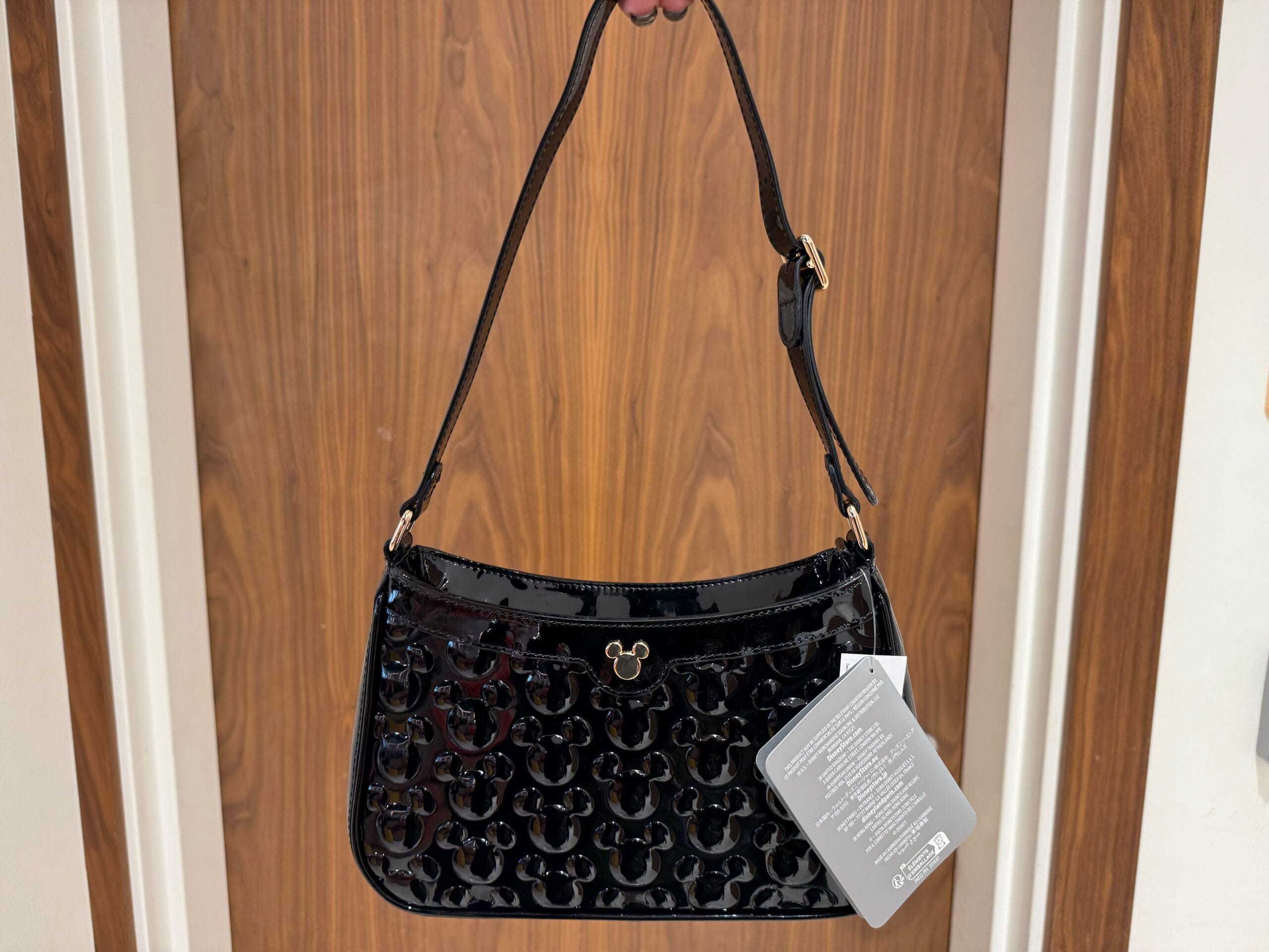 A black patent leather Icon Purse with a Mickey Mouse pattern and tag, held in front of a wooden door—classic Disney Parks style.