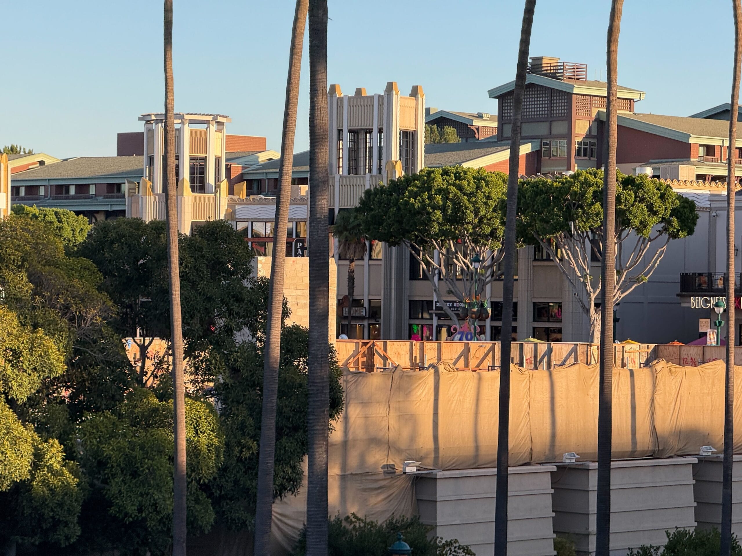 A building covered in tan construction tarps near Downtown Disney's shops and restaurants, trees, and a plaza at sunset. No castle visible.