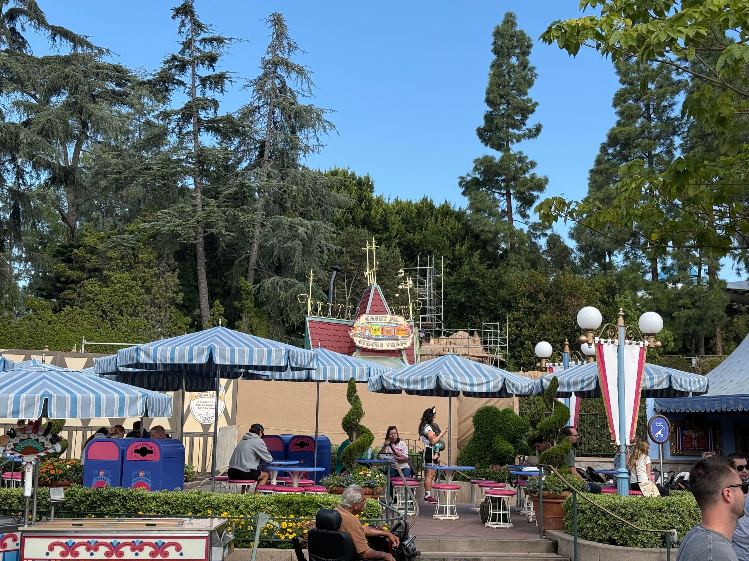 Guests relax at blue-striped umbrella tables by a construction zone in Disneyland’s Storybook Land, not far from Sleeping Beauty Castle.
