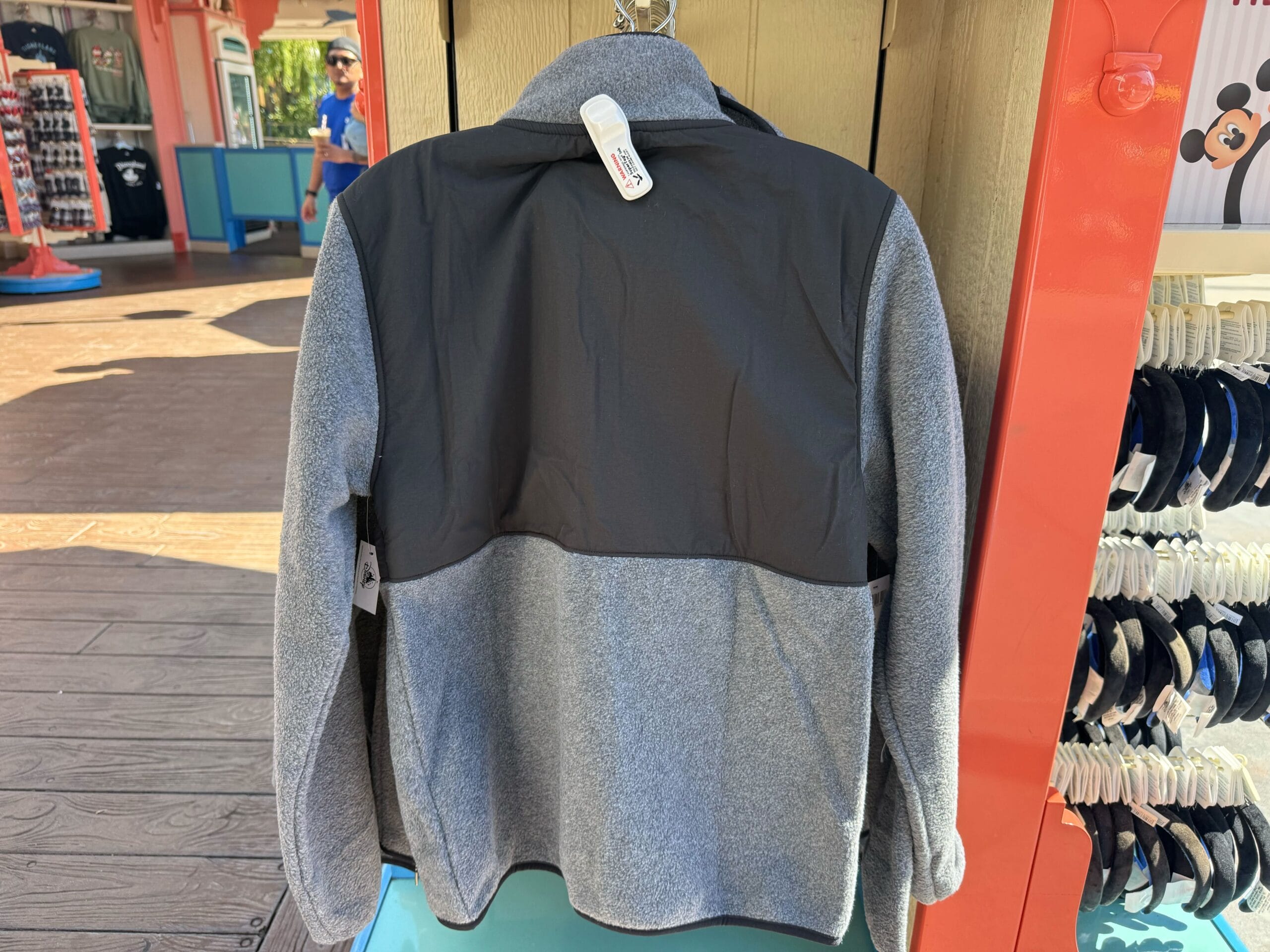 A gray and black jacket hangs on a rack in a Disney park shop, with themed hoodies and ear headbands displayed on the right.