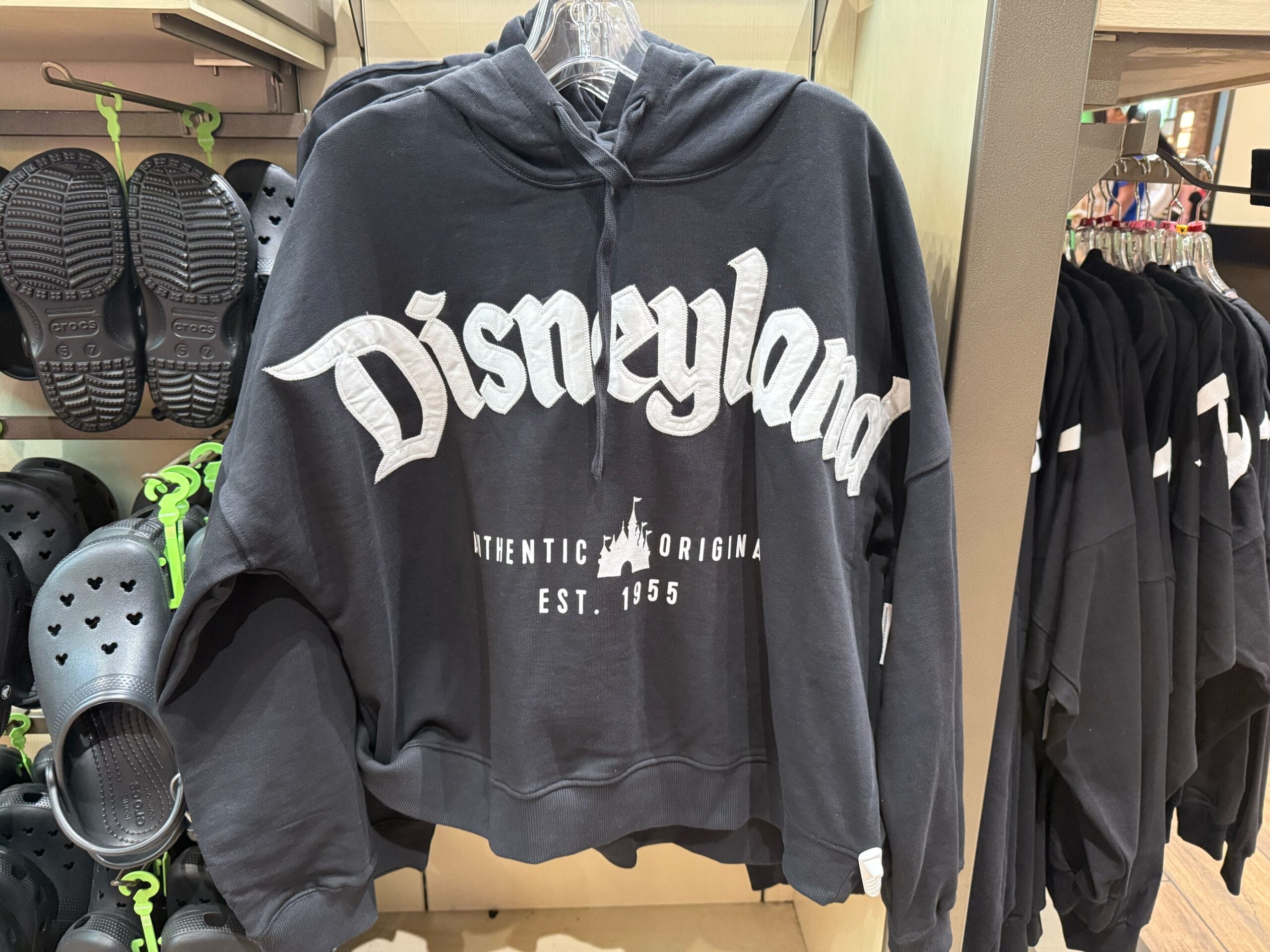Black Disneyland hoodie with white lettering, part of new arrivals, hangs by black Crocs—classic park merch, no castle in view.