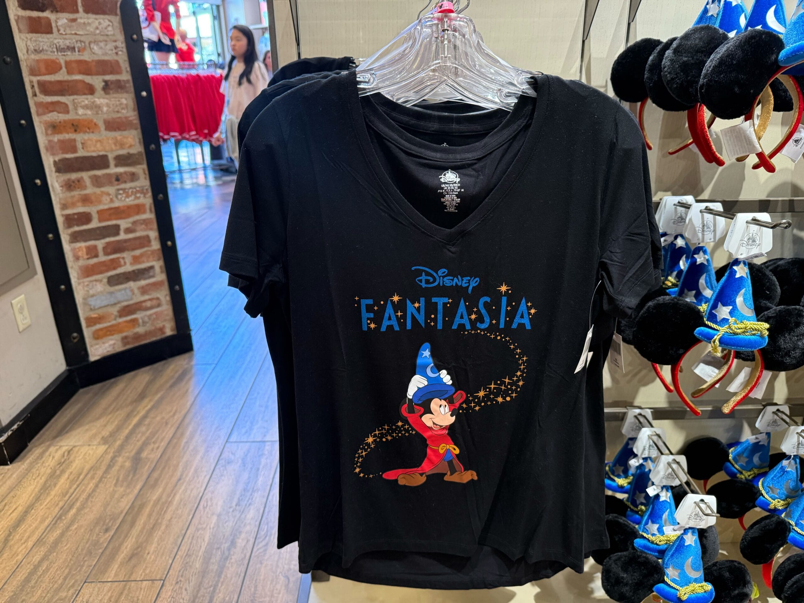 Black "Fantasia" tee with sorcerer Mickey, near mouse ear headbands and new arrivals at a Disney Park merchandise display.