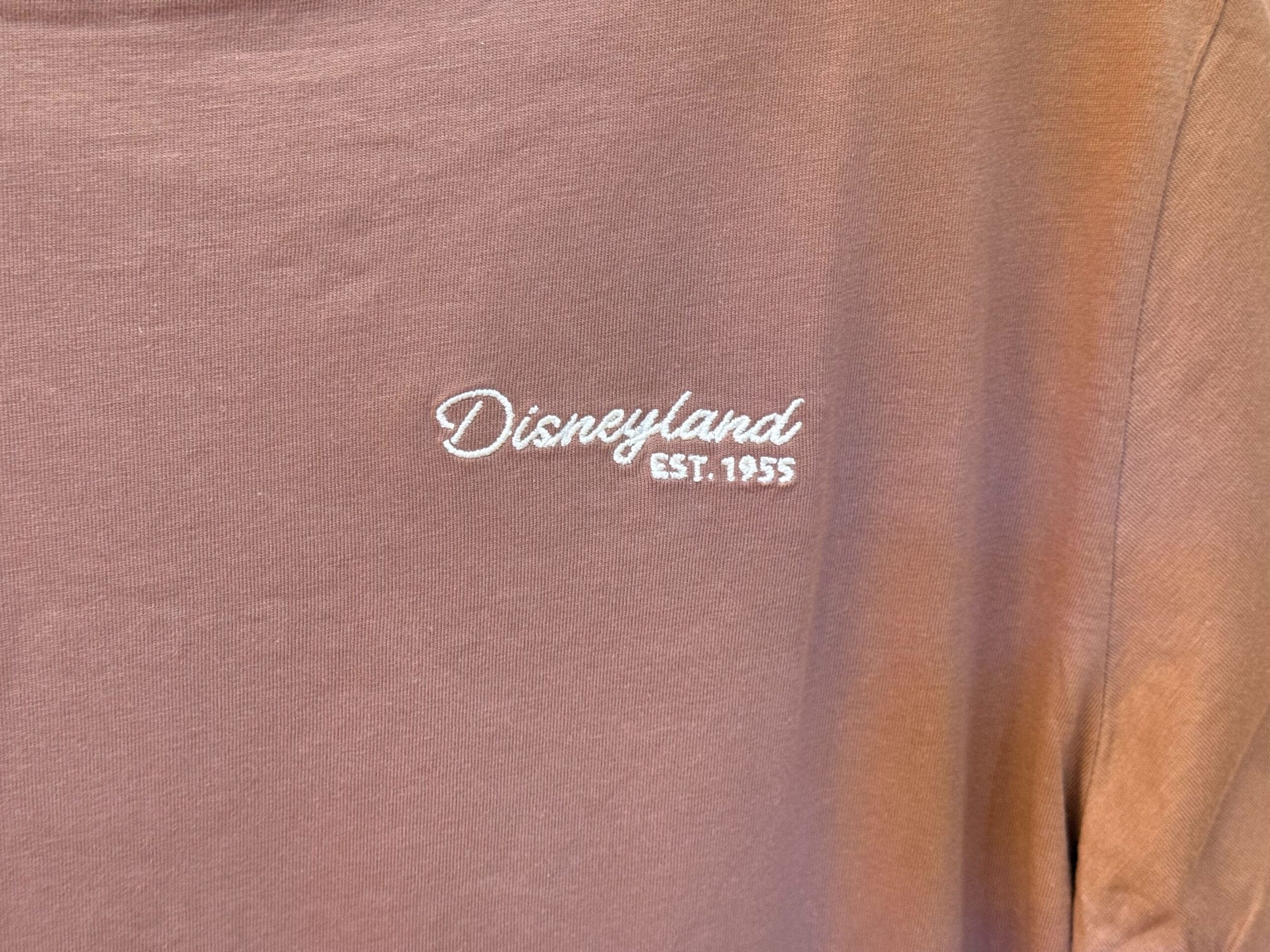 Detailed view of a brown Disneyland shirt, featuring white stitched "Disneyland EST. 1955," referencing the original Anaheim park.
