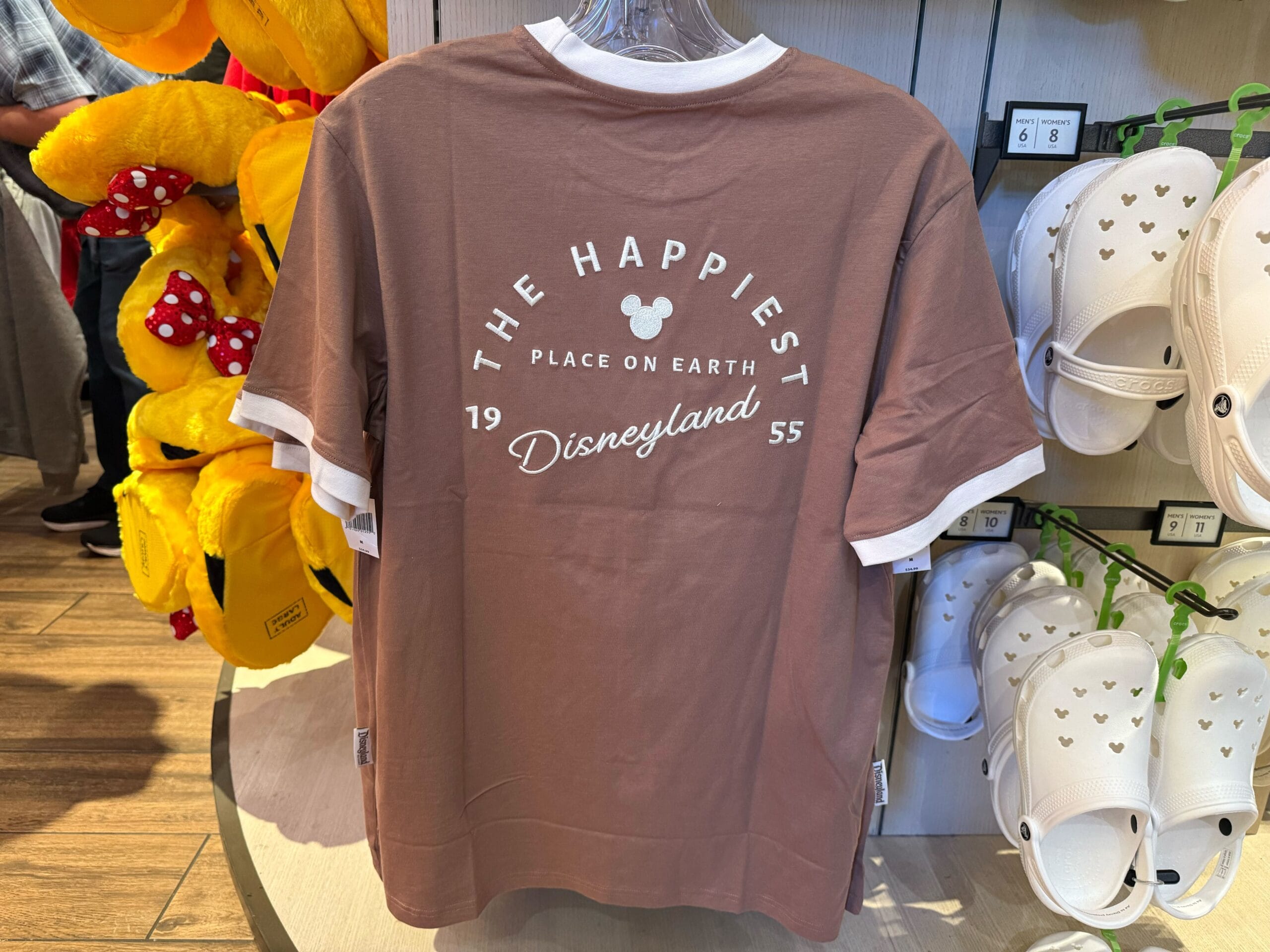 Brown Disneyland t-shirt with white text, "The Happiest Place on Earth, Disneyland 1955," seen among hoodies, referencing the classic Anaheim park—home to Sleeping Beauty Castle