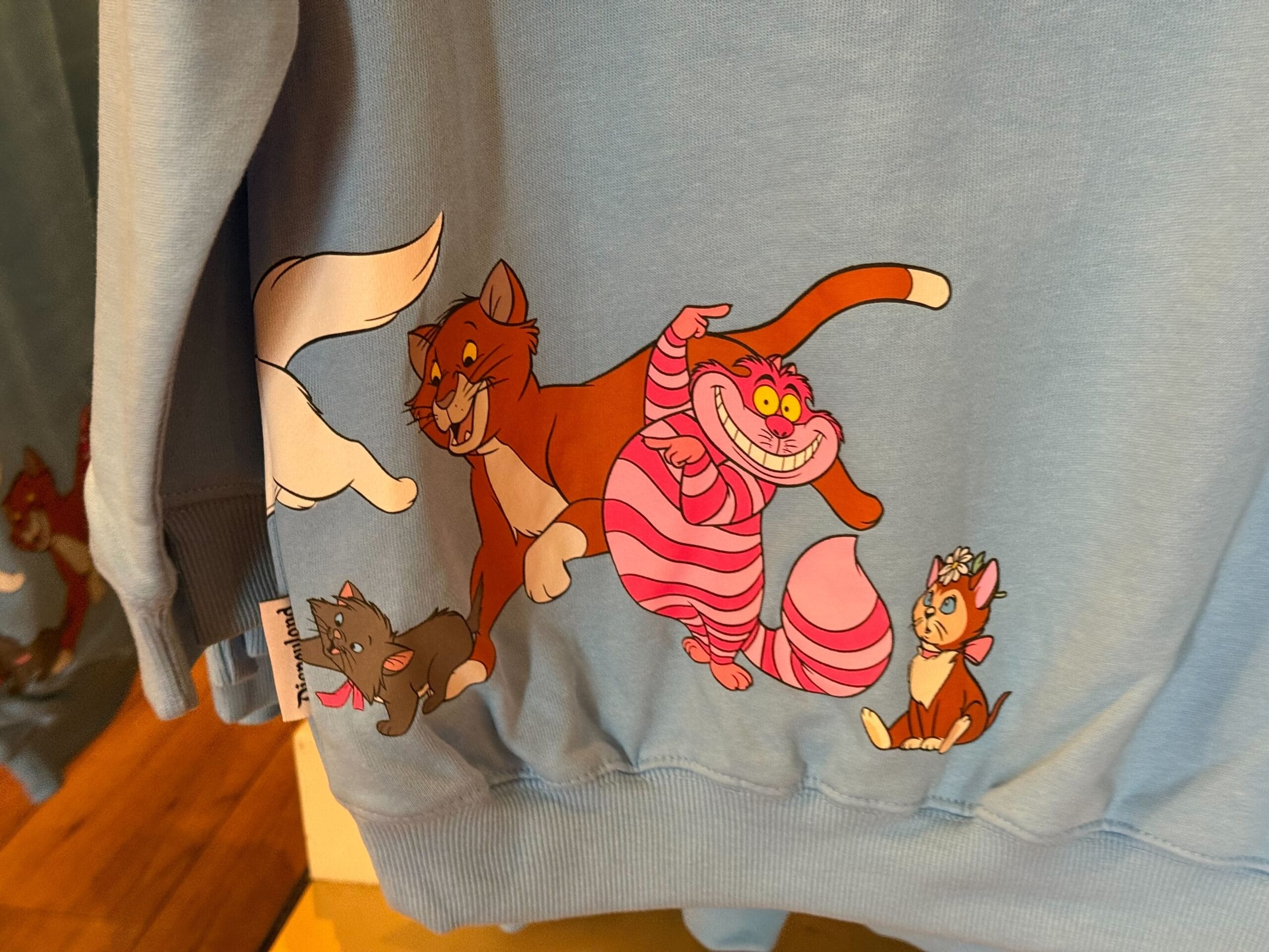Blue sweatshirt featuring cartoon cats, like a grinning pink-striped one and three playful friends, reminiscent of Disney merchandise.