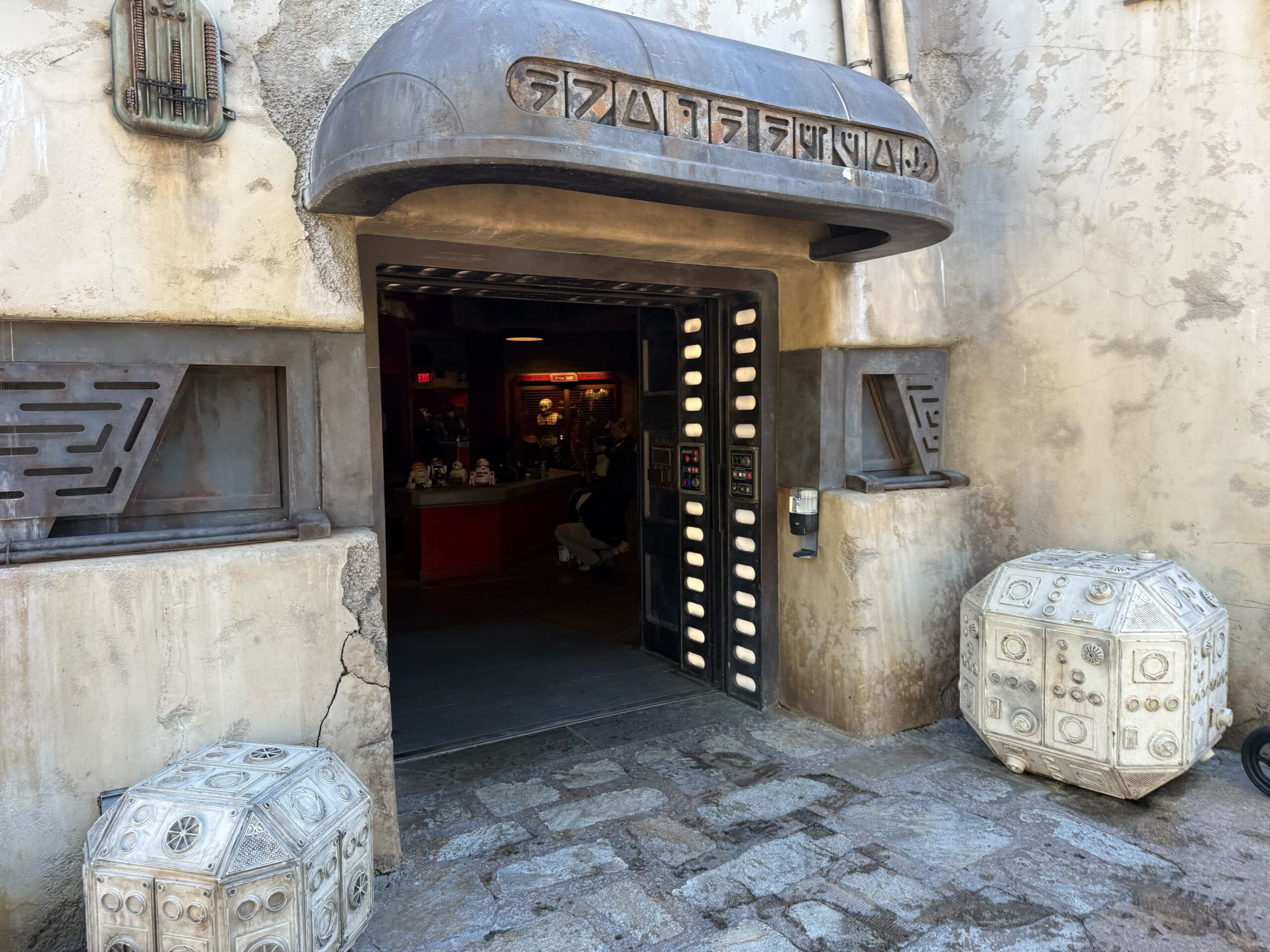 A sci-fi building entrance with alien script above the door, styled like Disneyland’s Droid Depot and featuring futuristic Disney flair.