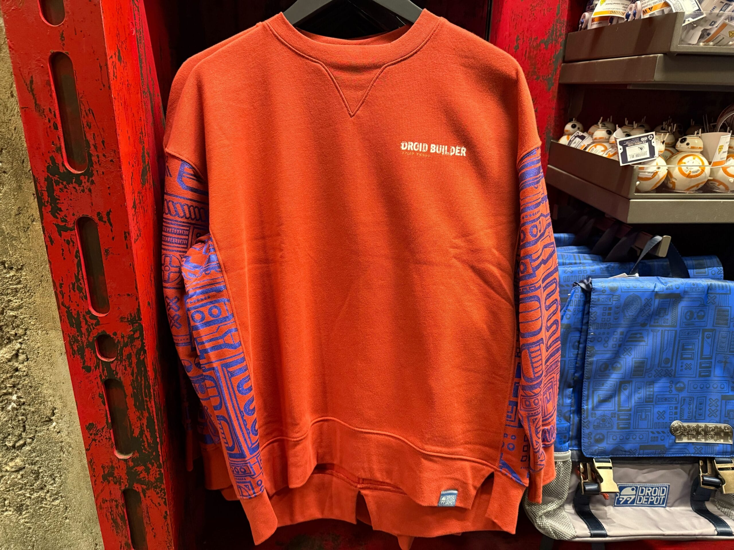 Red sweatshirt with blue patterned sleeves and "Droid Builder" text, exclusive Star Wars merch at Disneyland, near Sleeping Beauty Castle.