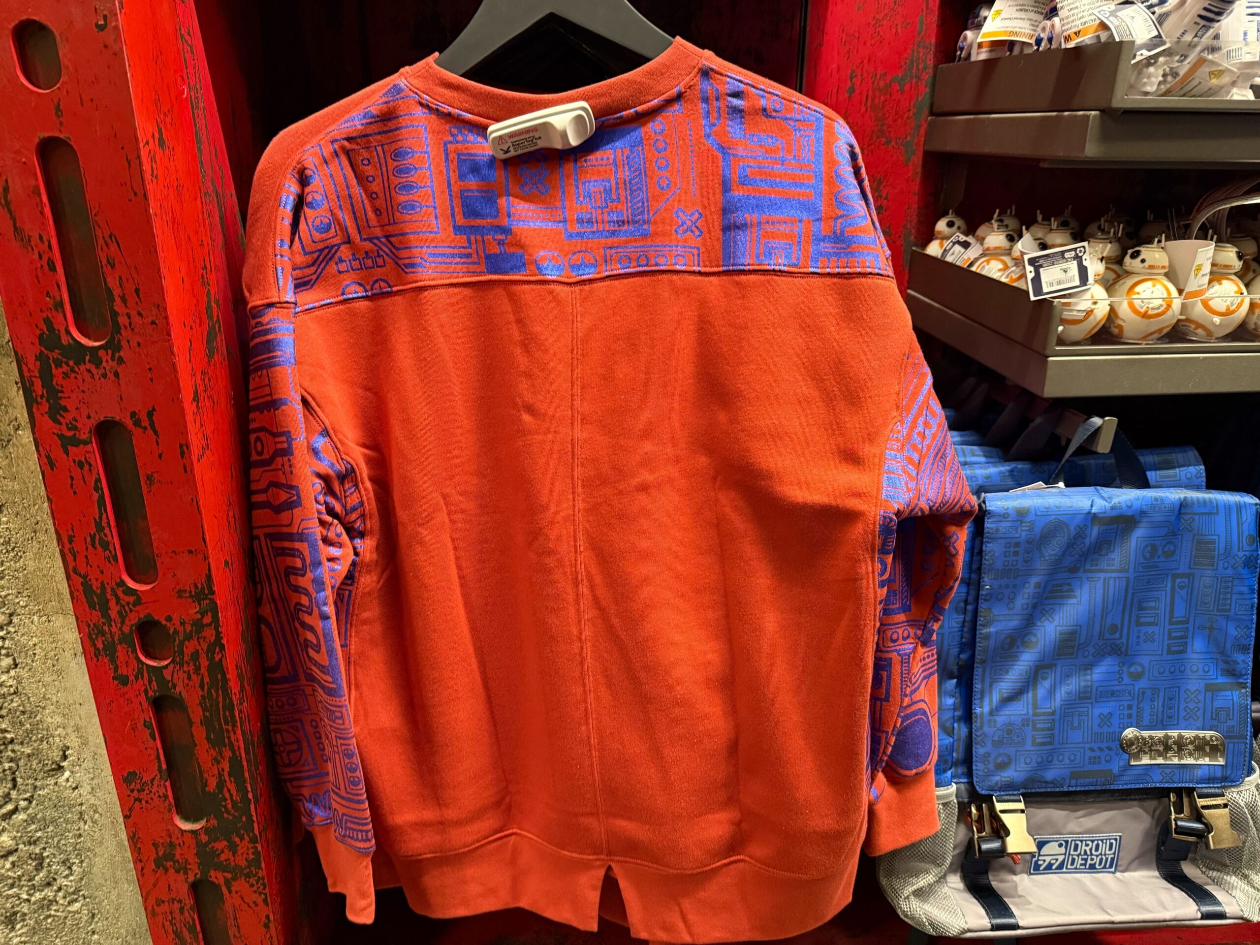 Red sweatshirt with blue geometric Star Wars patterns on display at Disneyland’s Droid Depot, surrounded by themed park merchandise.