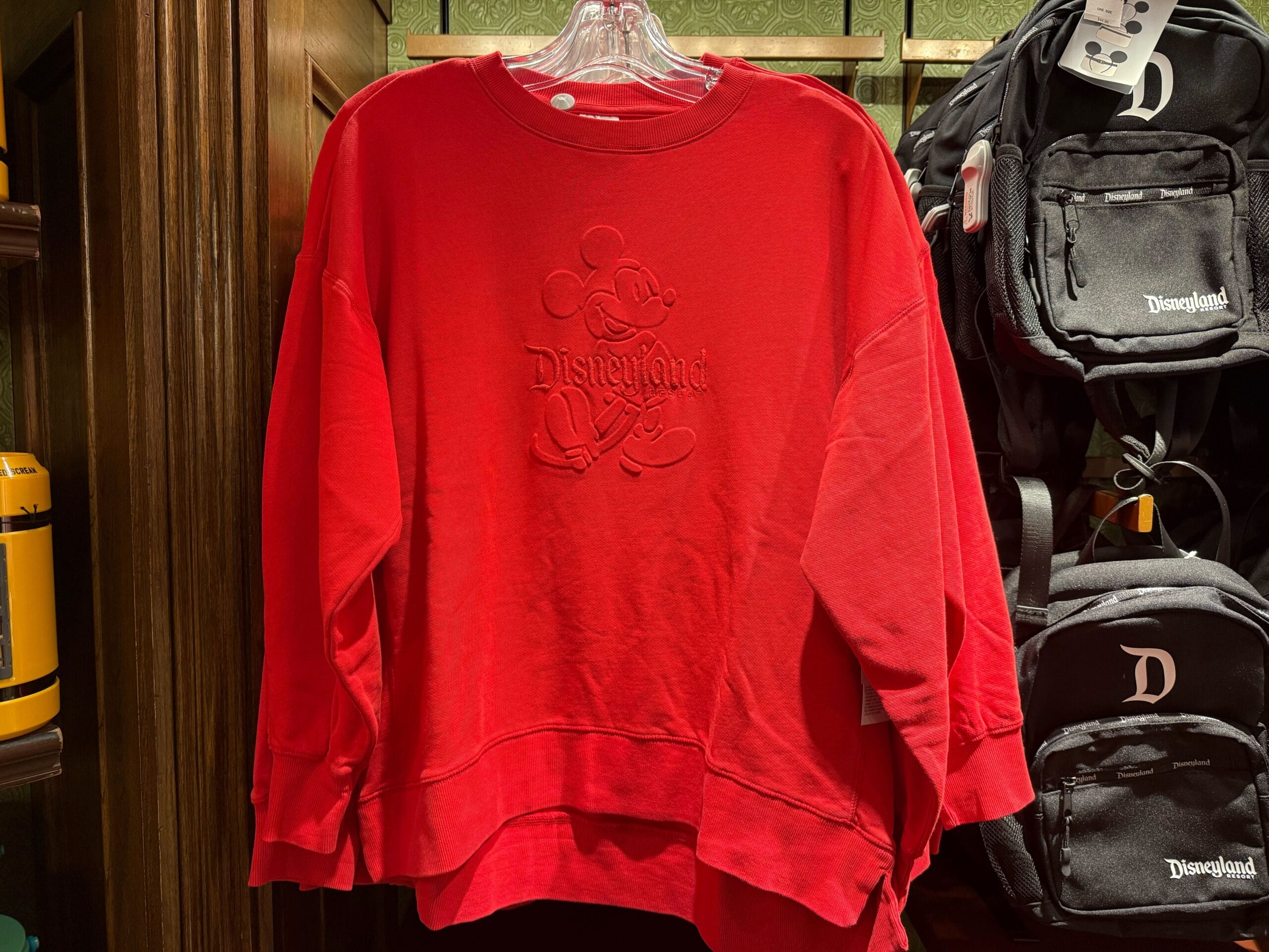 Red Disneyland sweatshirt with embossed Mickey, surrounded by black backpacks and park apparel; evokes classic Disney style.