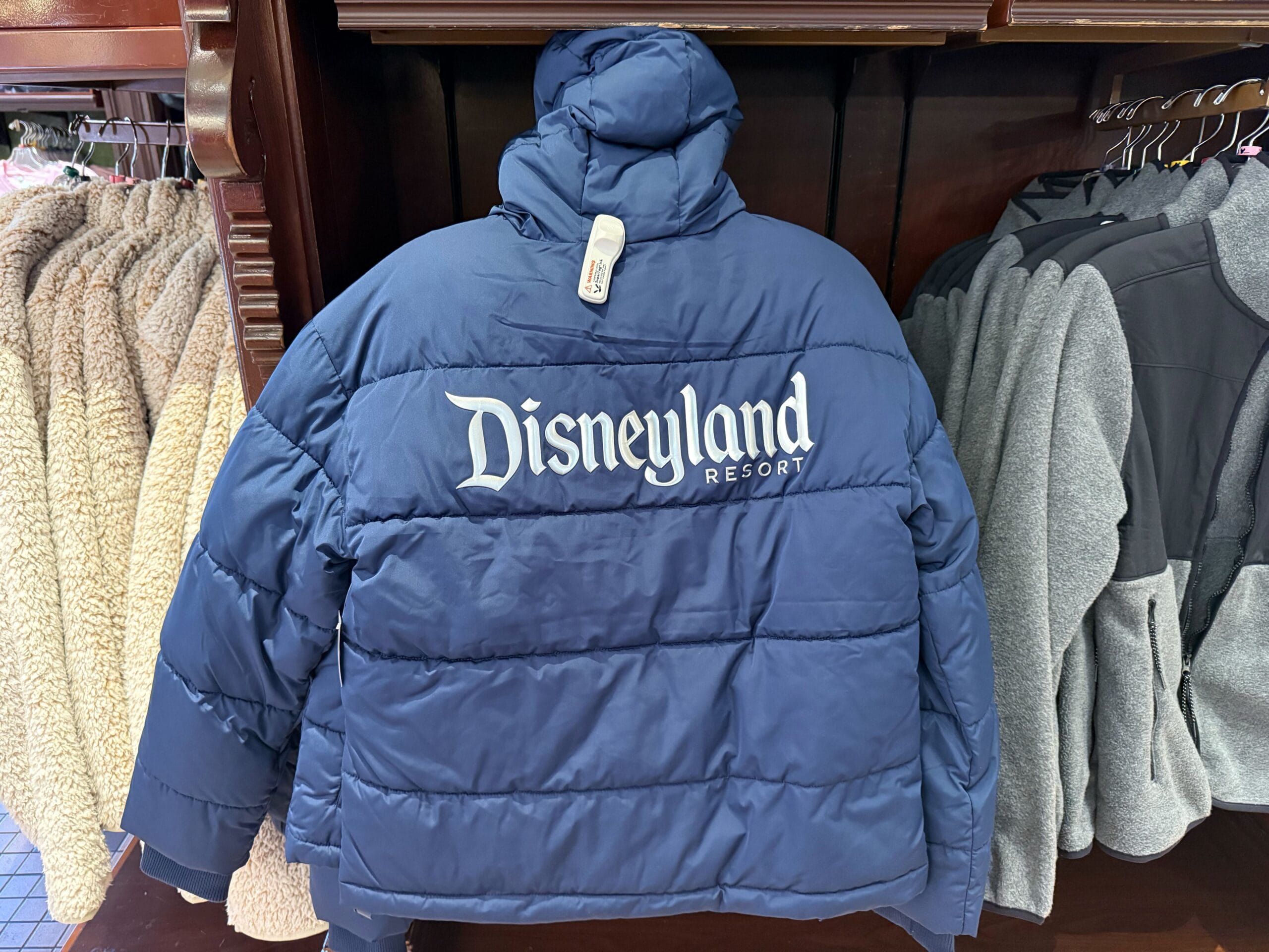 A blue puffer jacket with "Disneyland Resort" on the back, displayed among other park apparel, evokes classic Disneyland style.