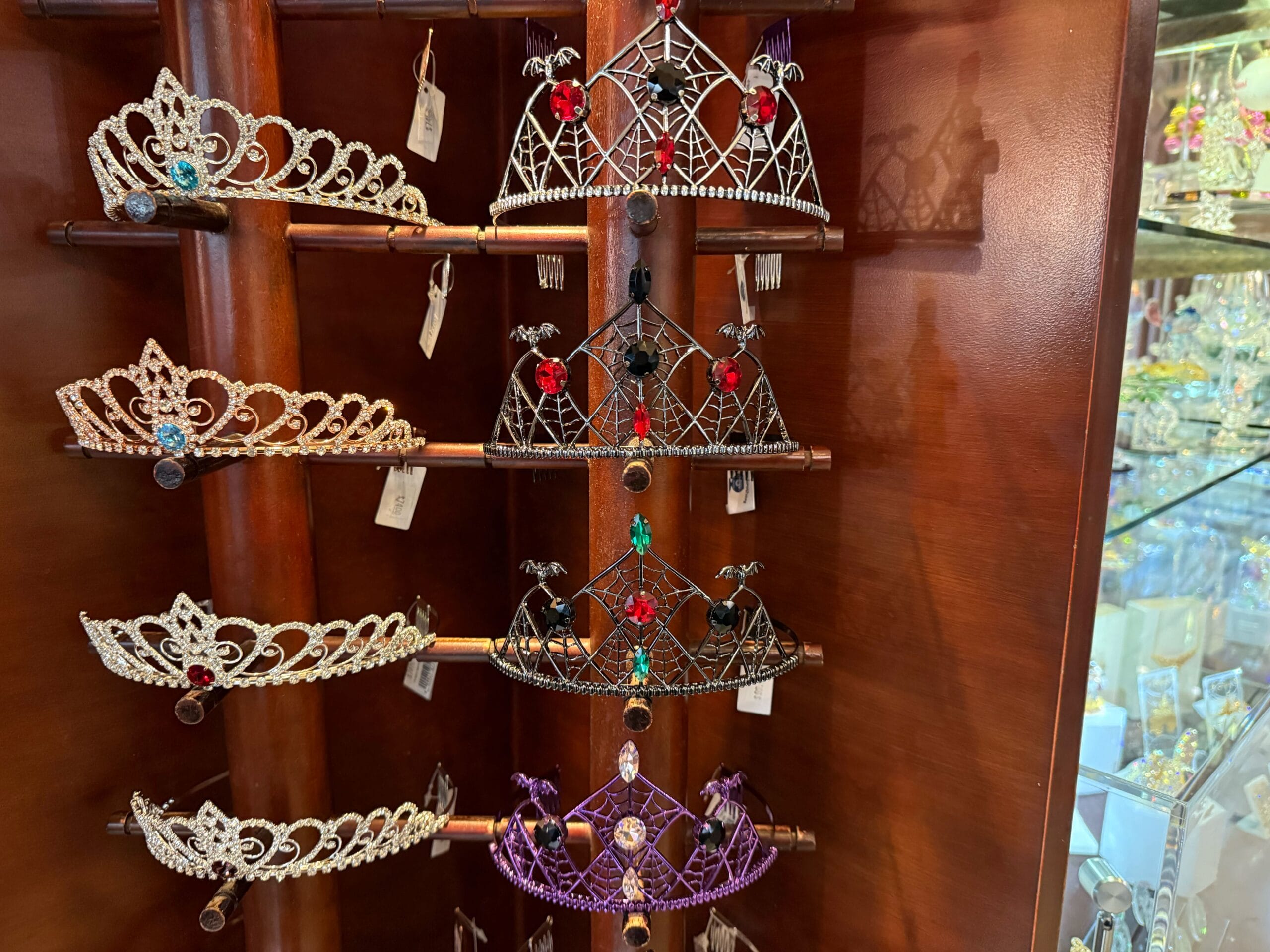 A collection of Halloween tiaras with vibrant gemstones on wooden shelves in a glass case, like Disney Parks' themed displays.
