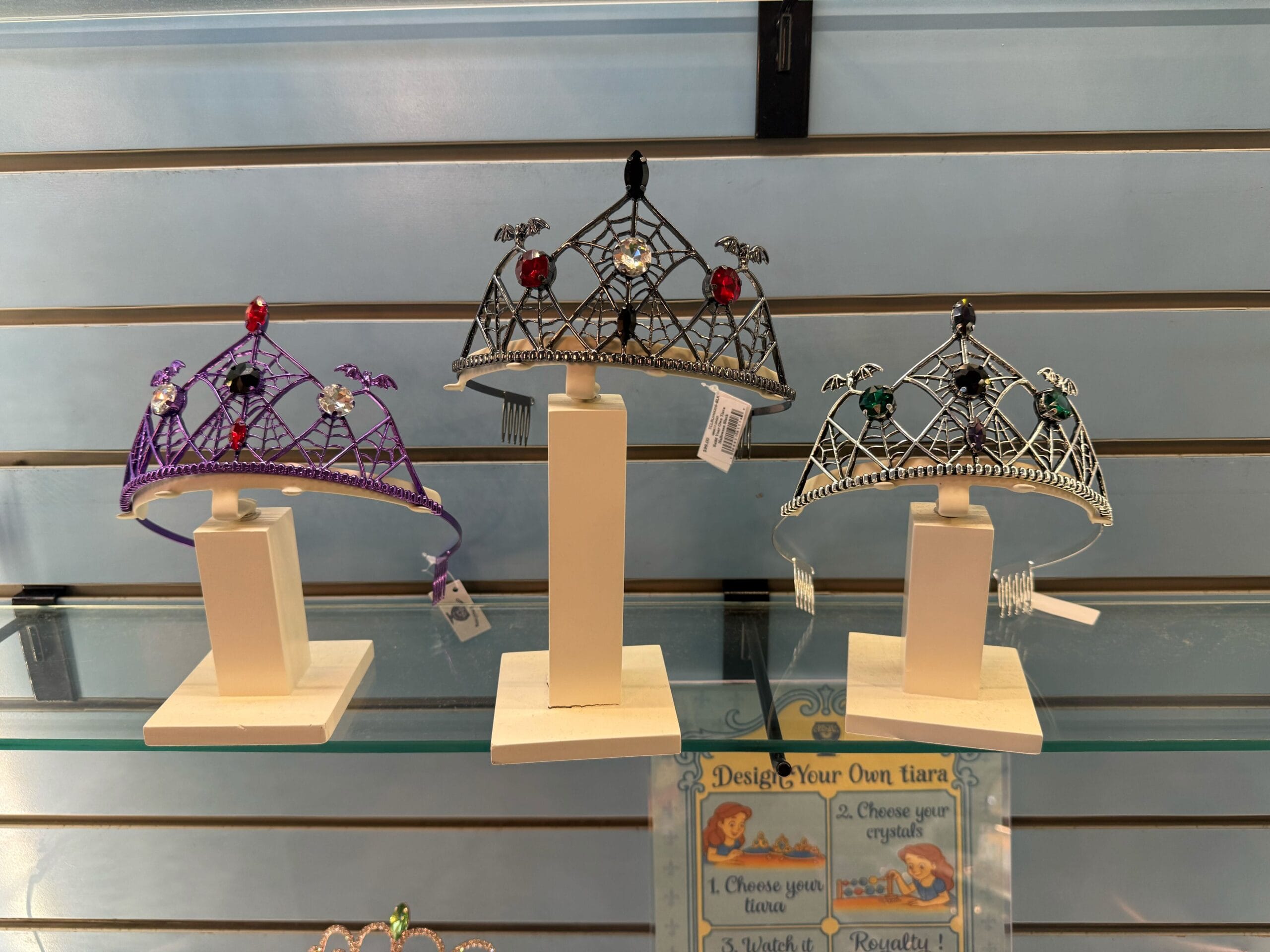 Three Halloween tiaras with colorful gems on stands, displayed like Disney Parks merchandise on a glass shelf, striped backdrop behind.