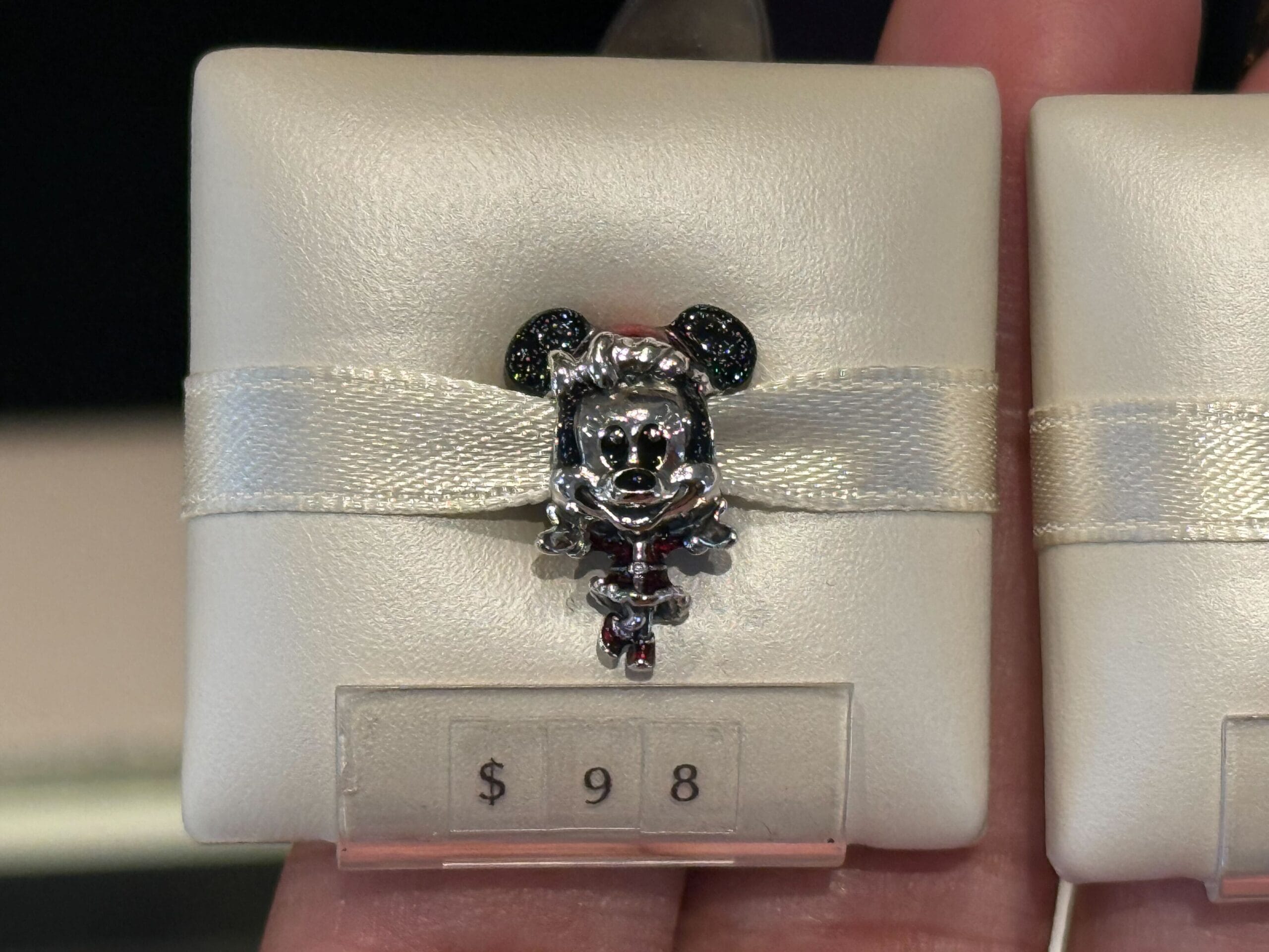 A silver Minnie Mouse Pandora charm with a bow, displayed on a white cushion in a Disney Park shop, priced at $98.