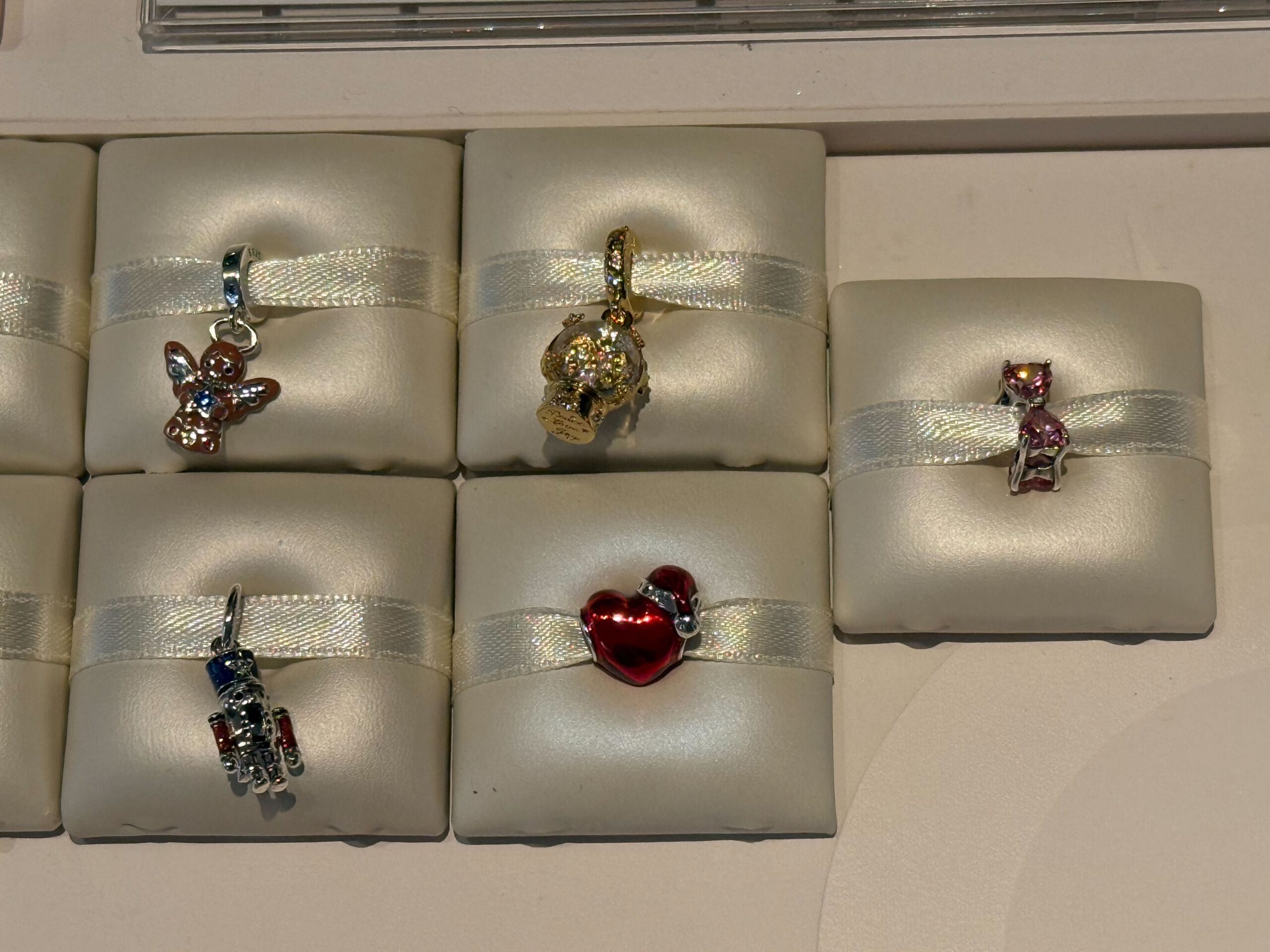 Five Pandora Charms on display, including a fairy and a bear reminiscent of Disney parks, set on cream-colored cushions.