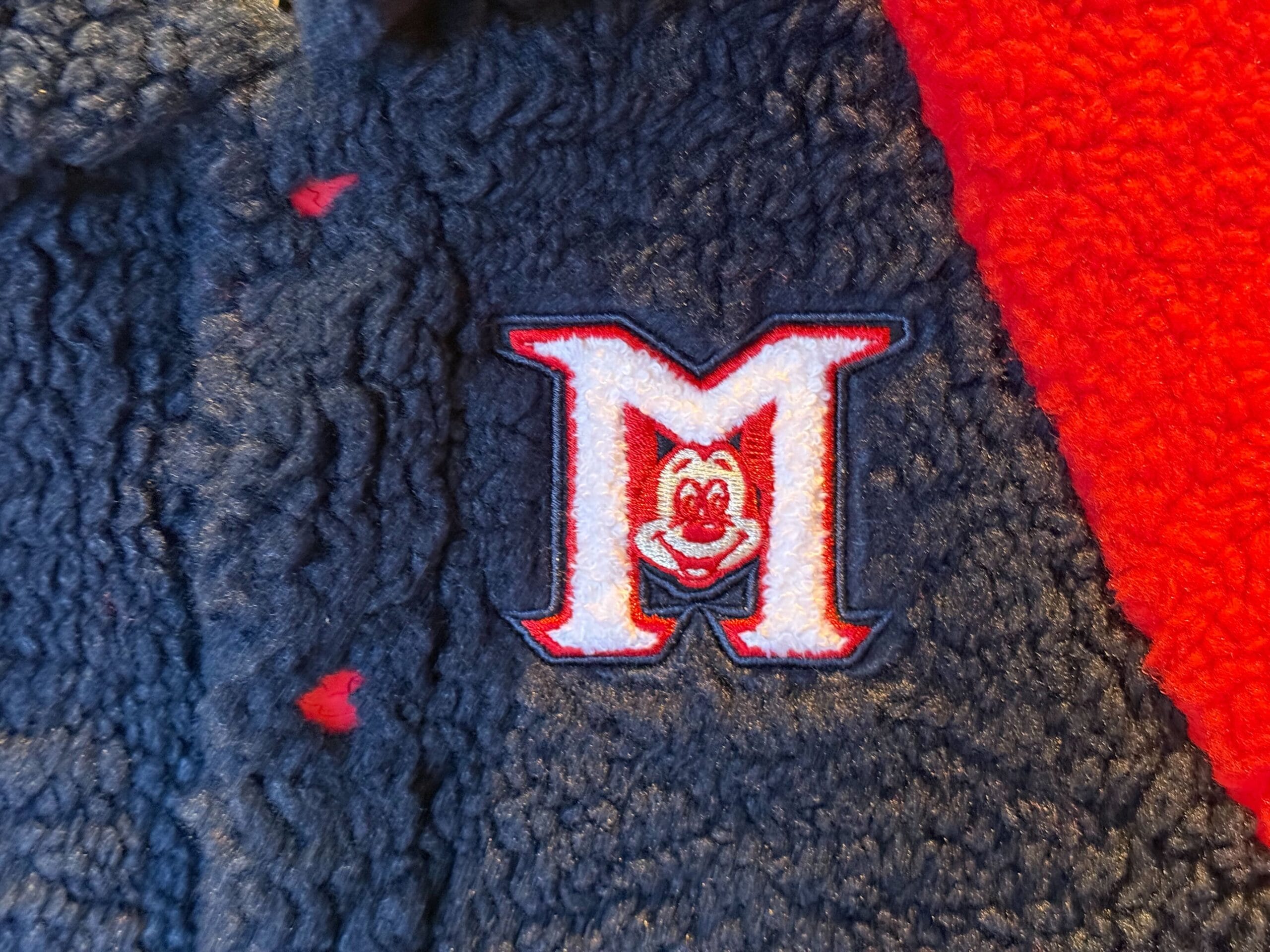 A red, white, and blue "M" patch with a cartoon face, inspired by Mickey Mouse Club merchandise on navy and red fleece.