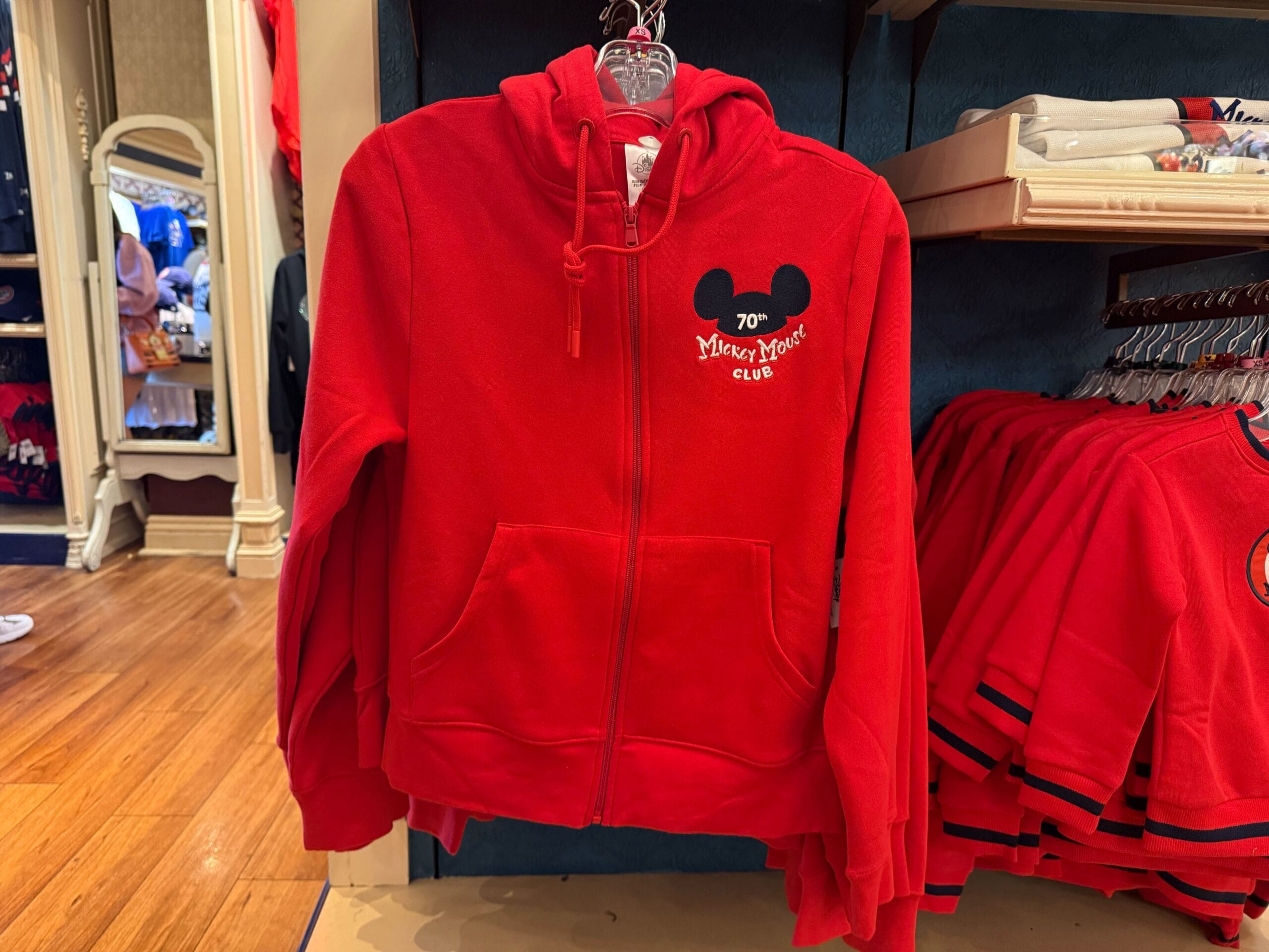 A red zip-up hoodie with a "70th Mickey Mouse Club" logo hangs on display as Disneyland merchandise in a store.