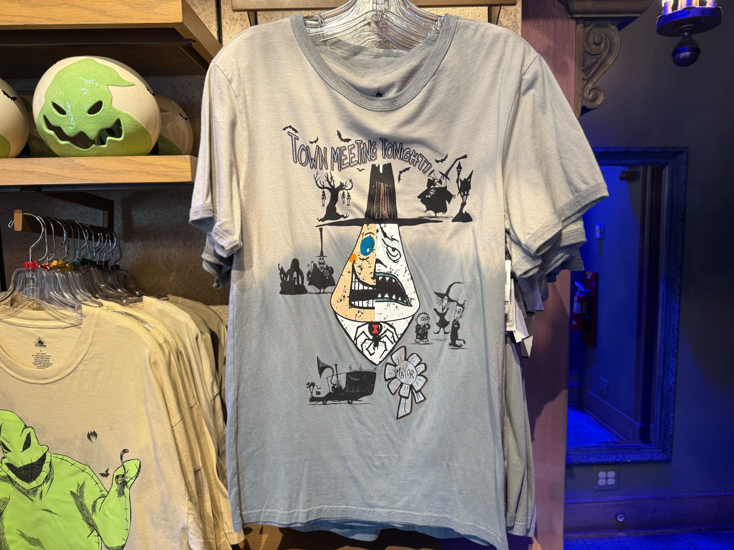Light gray tee in a park shop featuring "Town Meeting Tonight!!" plus Jack, Sally, and the Mayor from Nightmare Before Christmas.