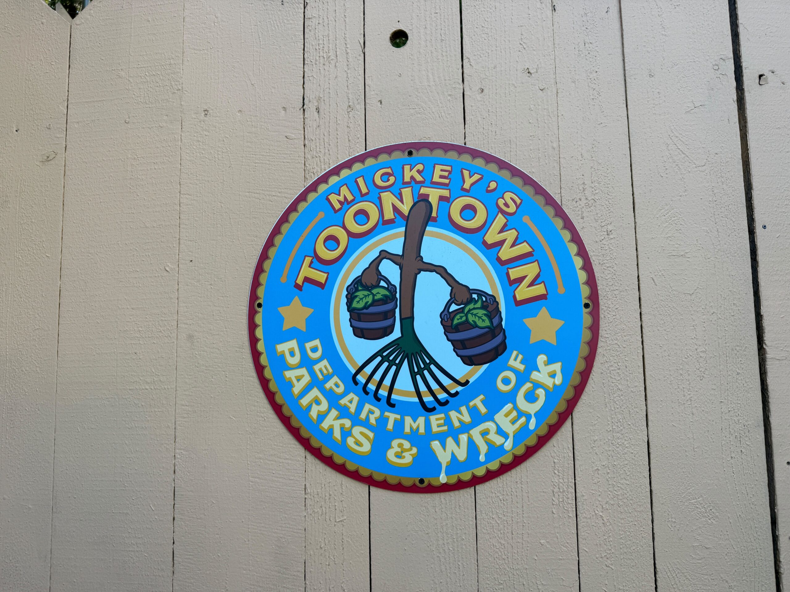 A round sign on a wooden fence in Mickey's Toontown at Disneyland says “Department of Parks & Wreck,” with rake and baskets.