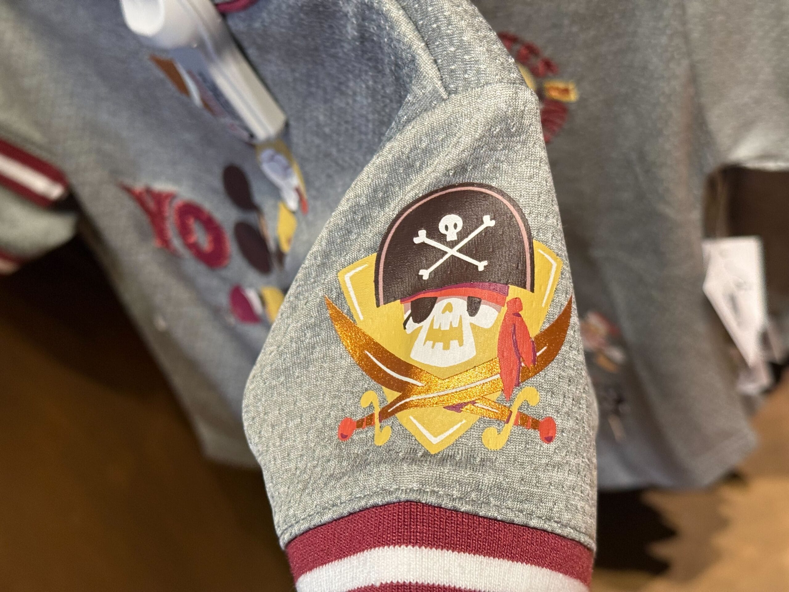 A pirate skull with hat, crossbones, swords, and red bird decorates a gray sleeve with maroon and white trim in Disney style.