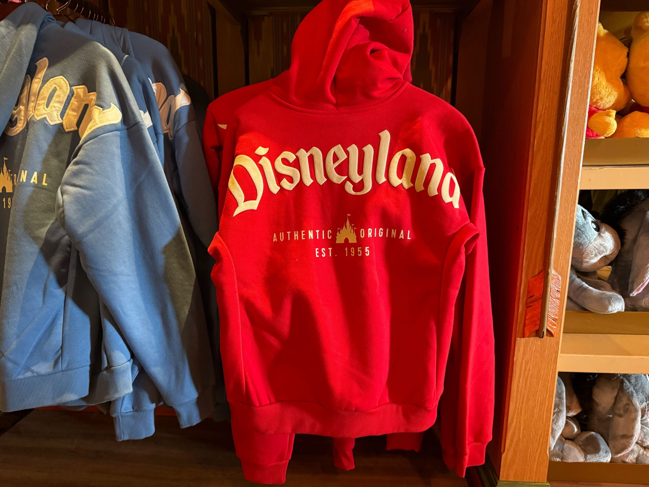 A red Disneyland hoodie with white lettering hangs on a rack beside a blue sweatshirt in a Disney Park store