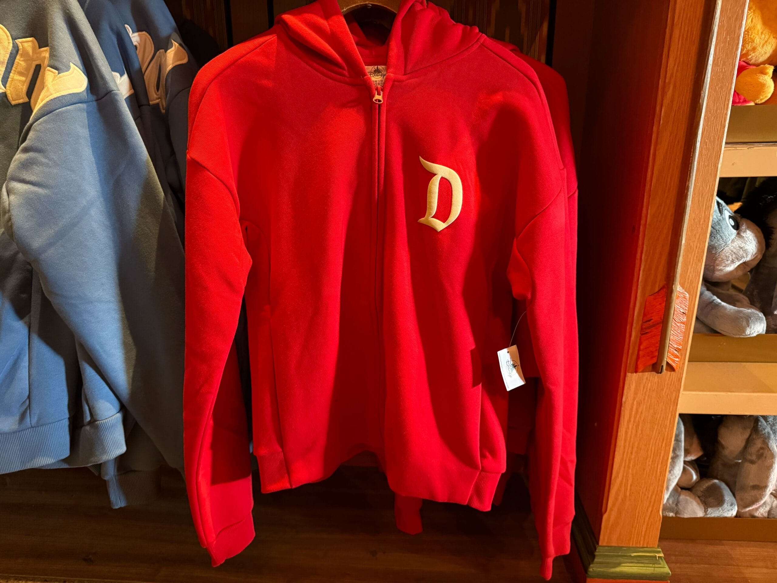 A red zip-up hoodie featuring a gold Disney "D" on the left chest, hanging near blue hoodies in a park gift shop display.