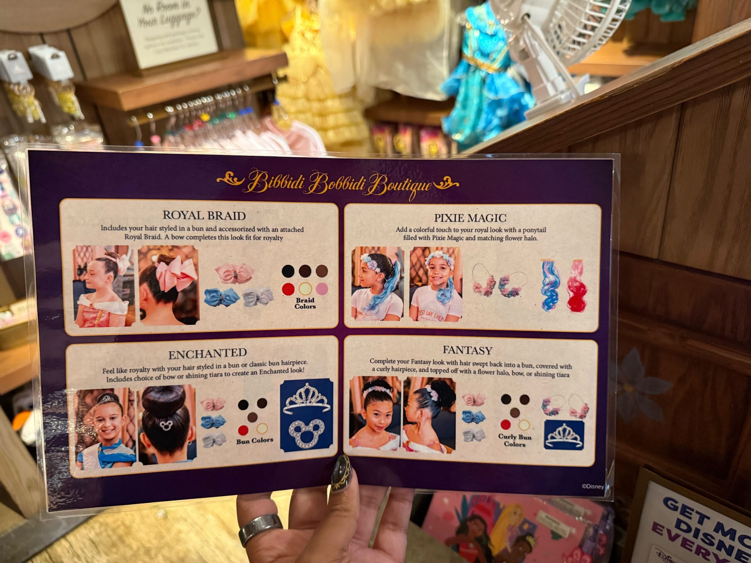 A person displays a Bibbidi Bobbidi Boutique sign showing four kids’ hairstyle choices, with photos like those found at Disney parks.