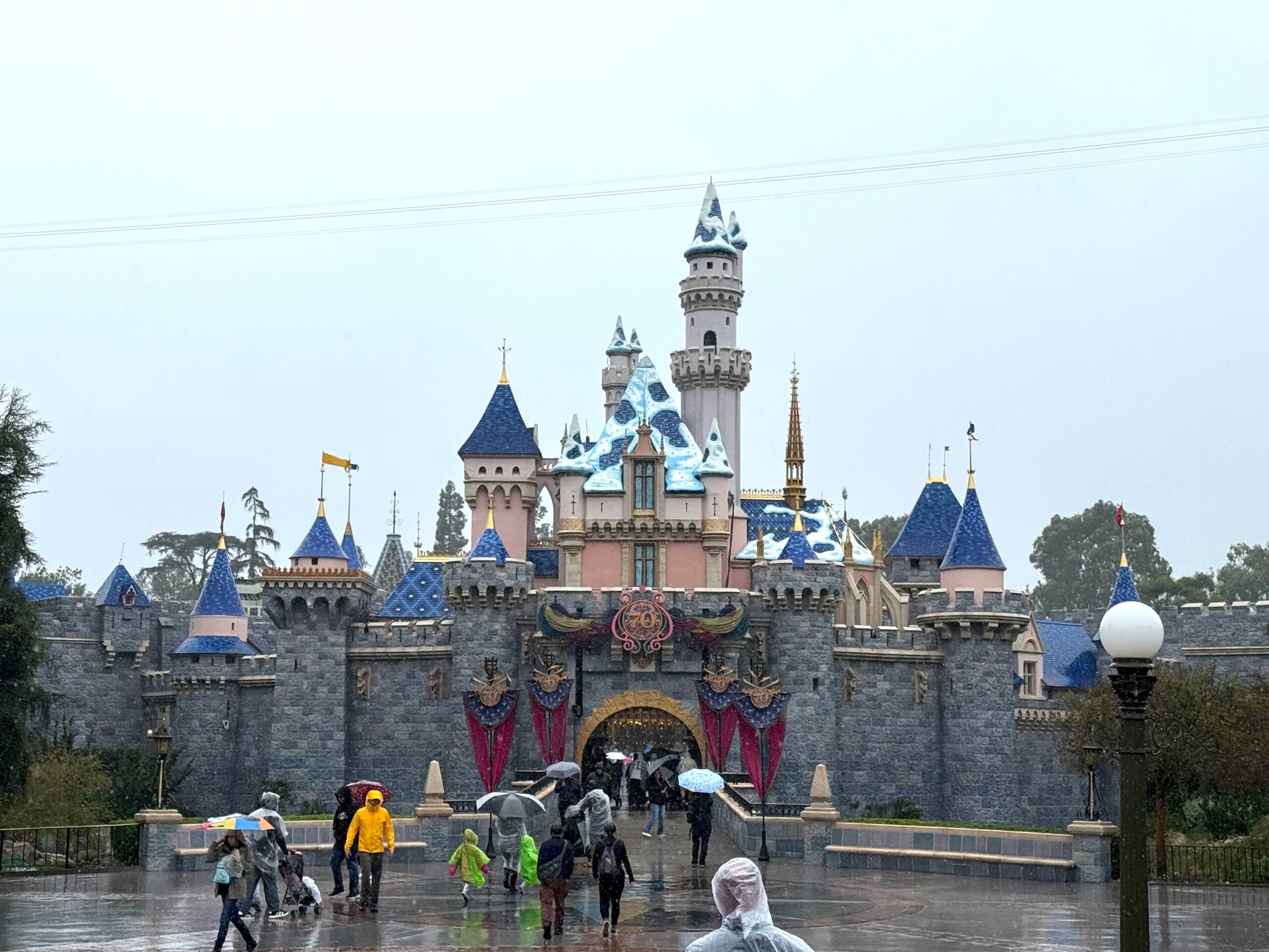 Disneyland’s Sleeping Beauty Castle on a rainy day, smaller than Walt Disney World’s Cinderella Castle, guests with umbrellas in front.