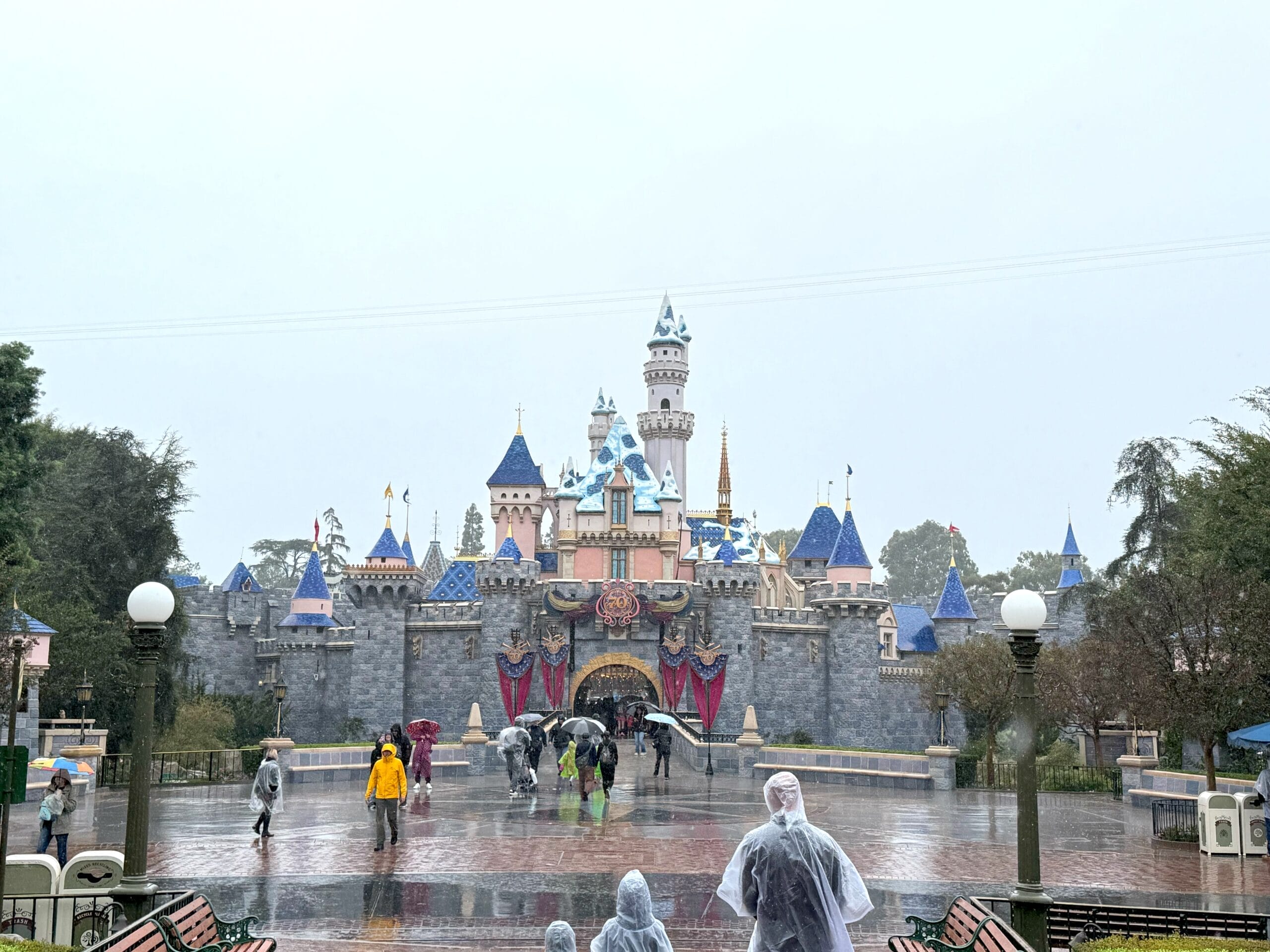 Guests in raincoats walk along Main Street toward Disneyland’s  pink and blue Sleeping Beauty Castle, which already has snow atop its turrets.