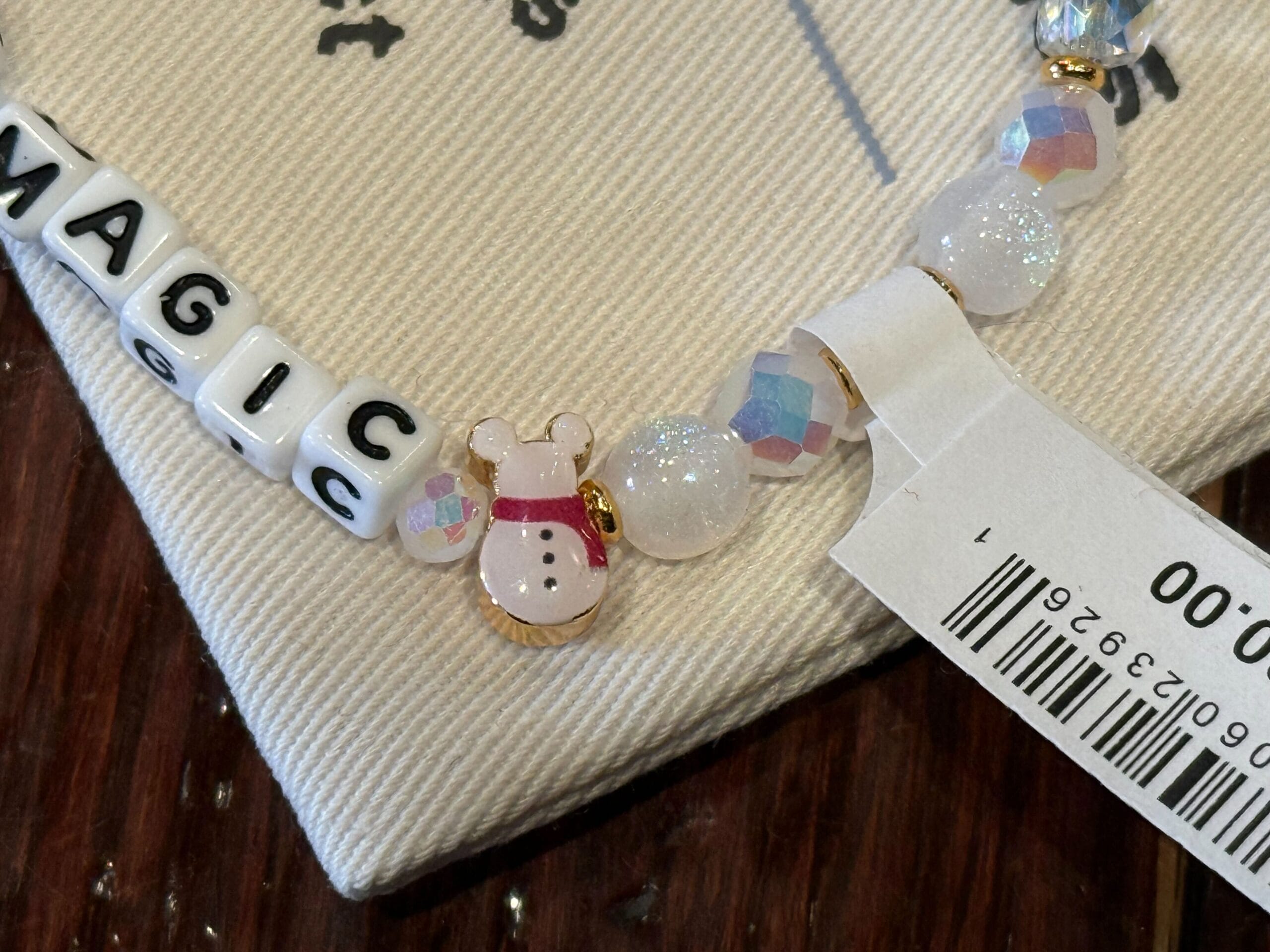 Beaded bracelet features white "MAGIC" letter beads and a snowman charm, styled after 2025 Disneyland holiday merch. No castle visible.
