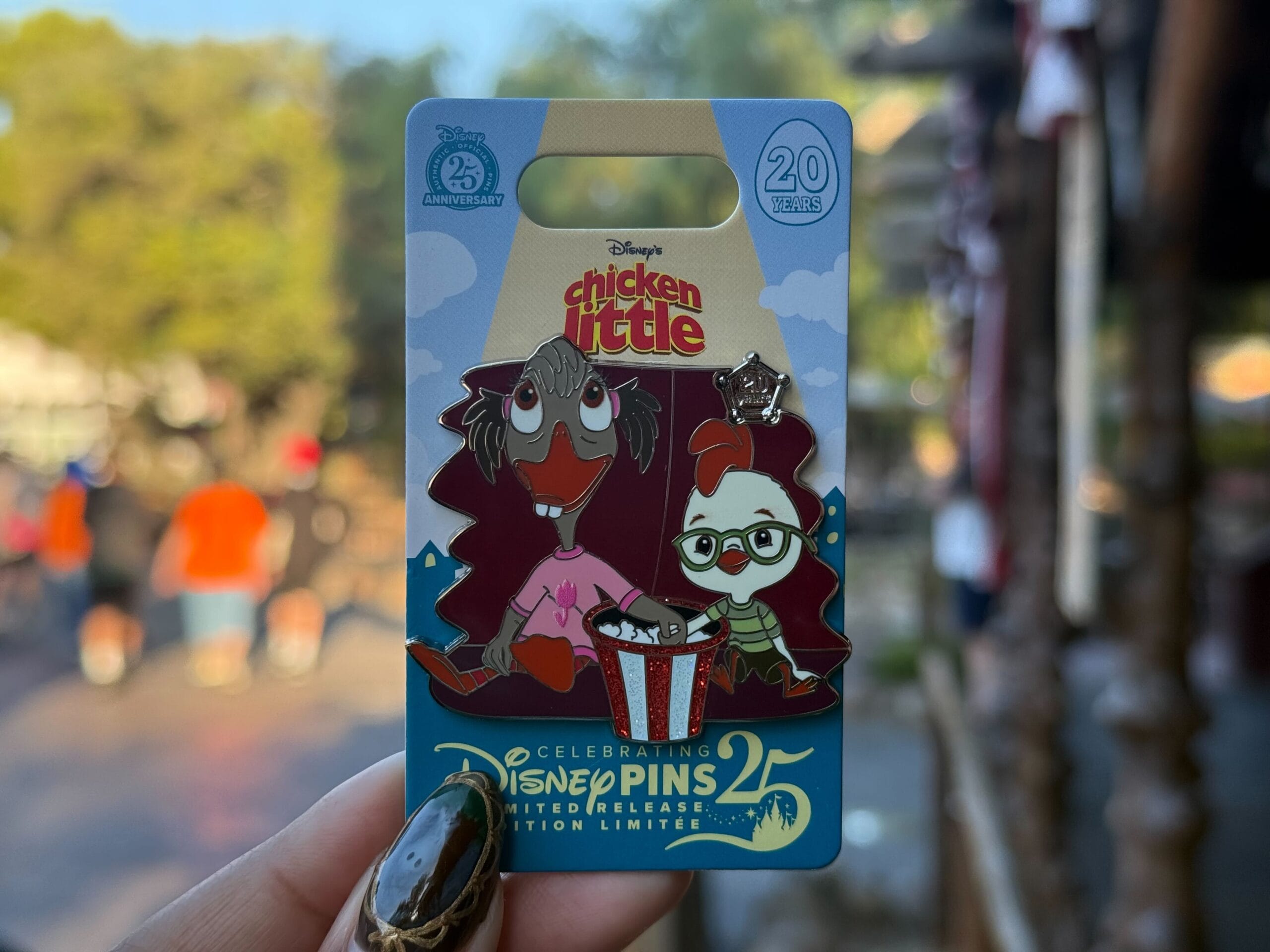 A hand displays a Disneyland Chicken Little 25th anniversary pin, held up outdoors with a blurred Disney park background behind it.