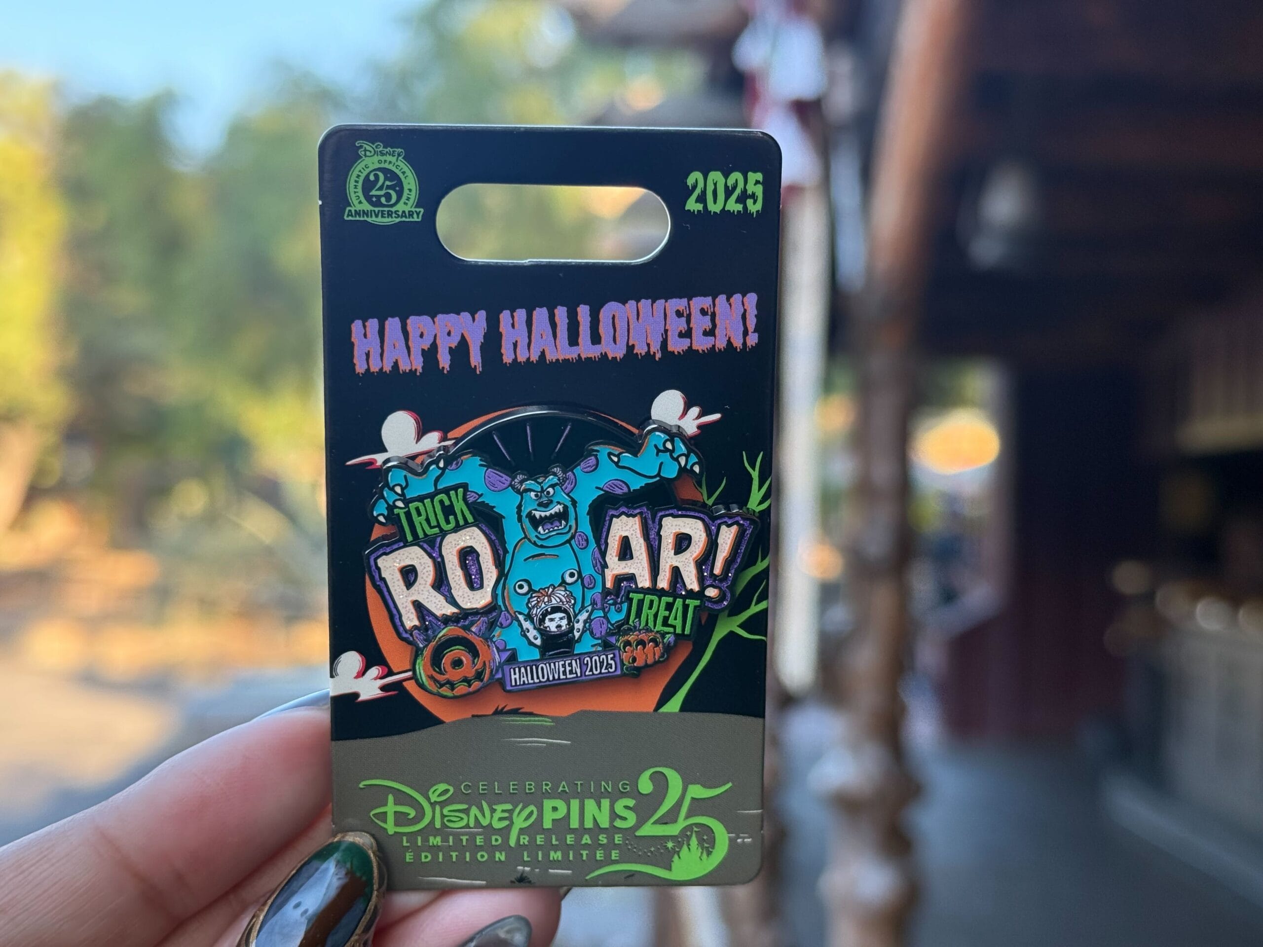 A hand holds a Disneyland 25th anniversary Halloween pin featuring Sulley from Monsters, Inc. and "ROAR! Trick or Treat" text.