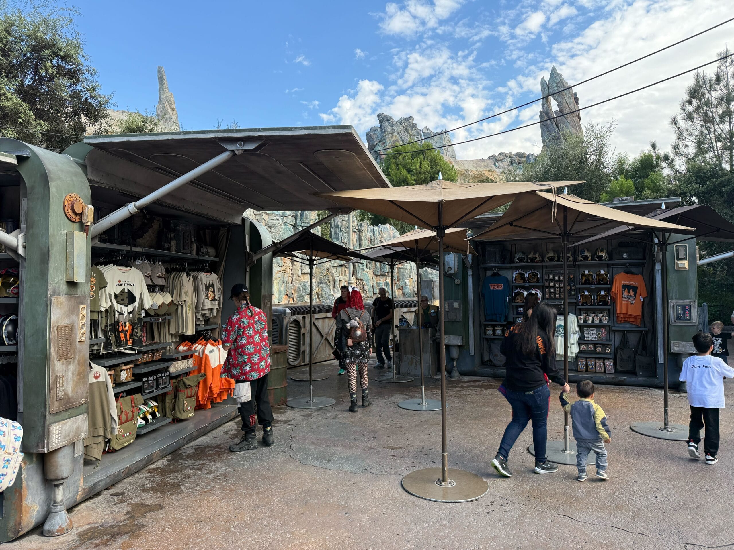 Sci-fi market stalls at a Disney park offer Star Wars goods as guests stroll with umbrellas, reminiscent of Galaxy’s Edge.