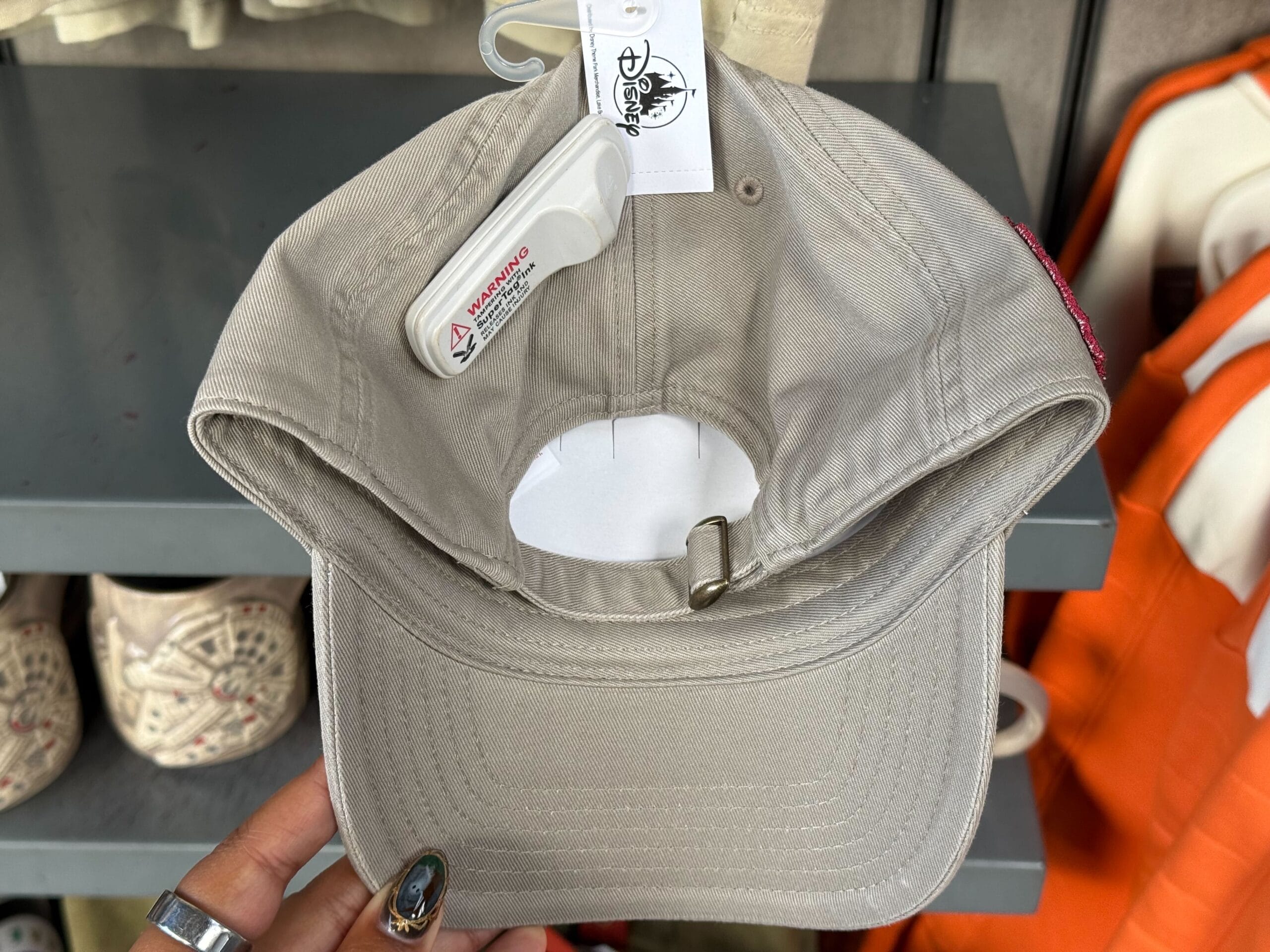 A hand displays a beige baseball cap with a security tag in a Disneyland shop, surrounded by other Disney hats and apparel.