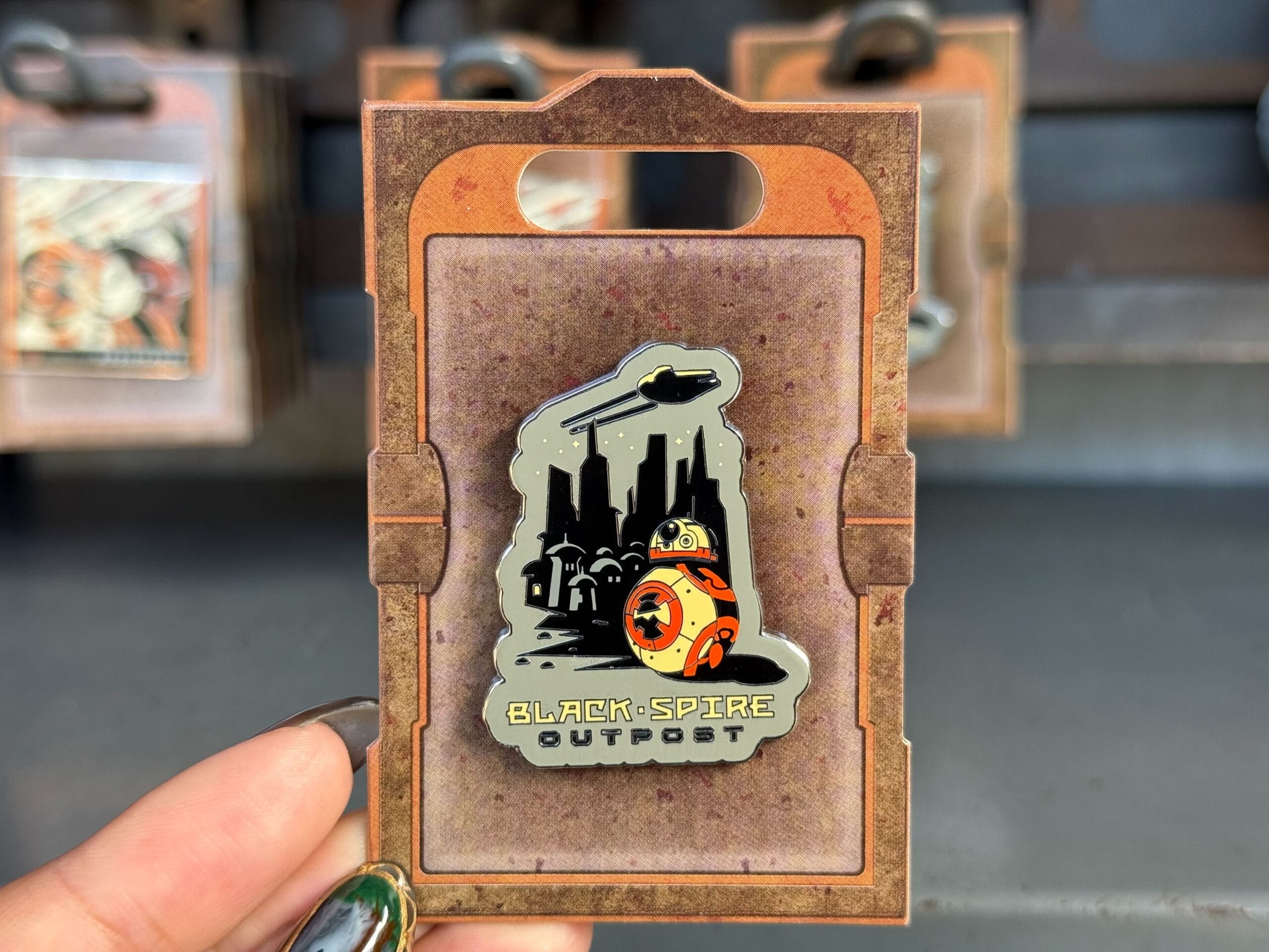 A hand shows a "Black Spire Outpost" pin with BB-8 and spaceships at Disneyland, home to the original Sleeping Beauty Castle.