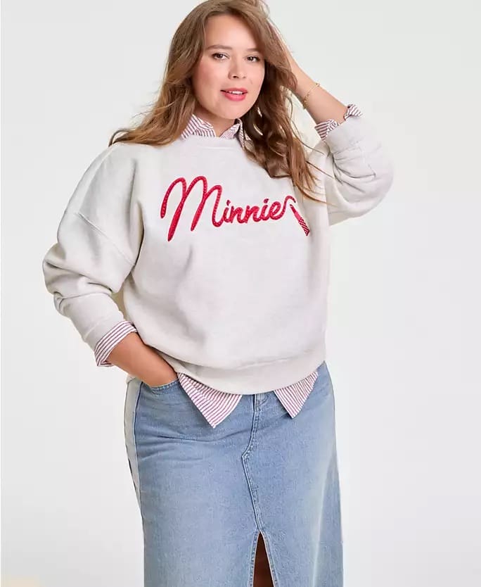 Woman in a white "Minnie" sweatshirt and denim skirt, Disney 2025 Holiday Collection, posing casually at a Disney park.