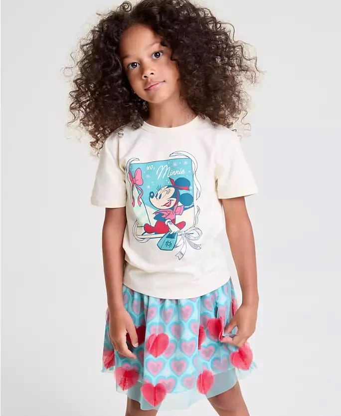Young girl in Minnie Mouse tee and blue skirt from 2025 Disney Holiday Collection, smiling at a Disney park, no castle visible.