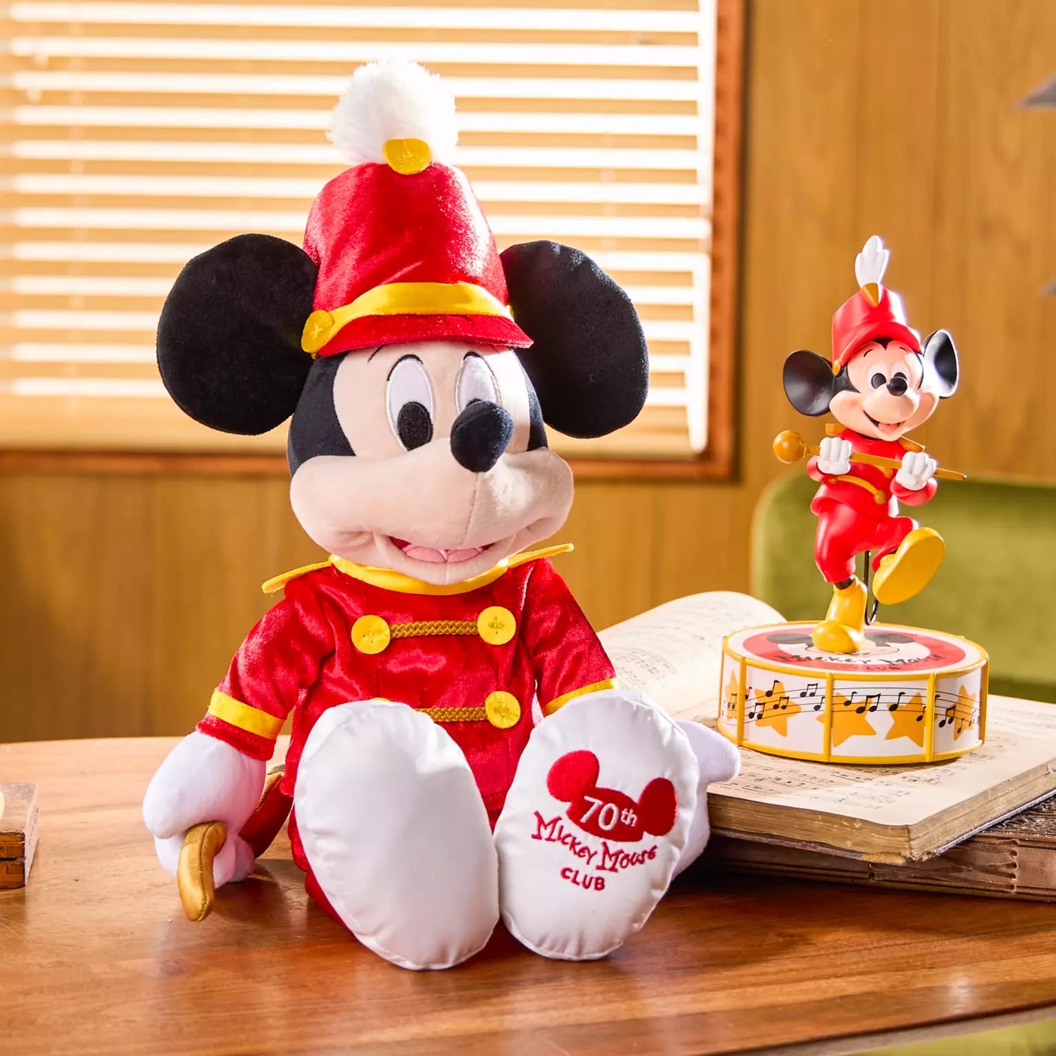 Disney Store-Exclusive plush and figurine of Mickey Mouse in a red band uniform on a desk with wooden blinds behind.