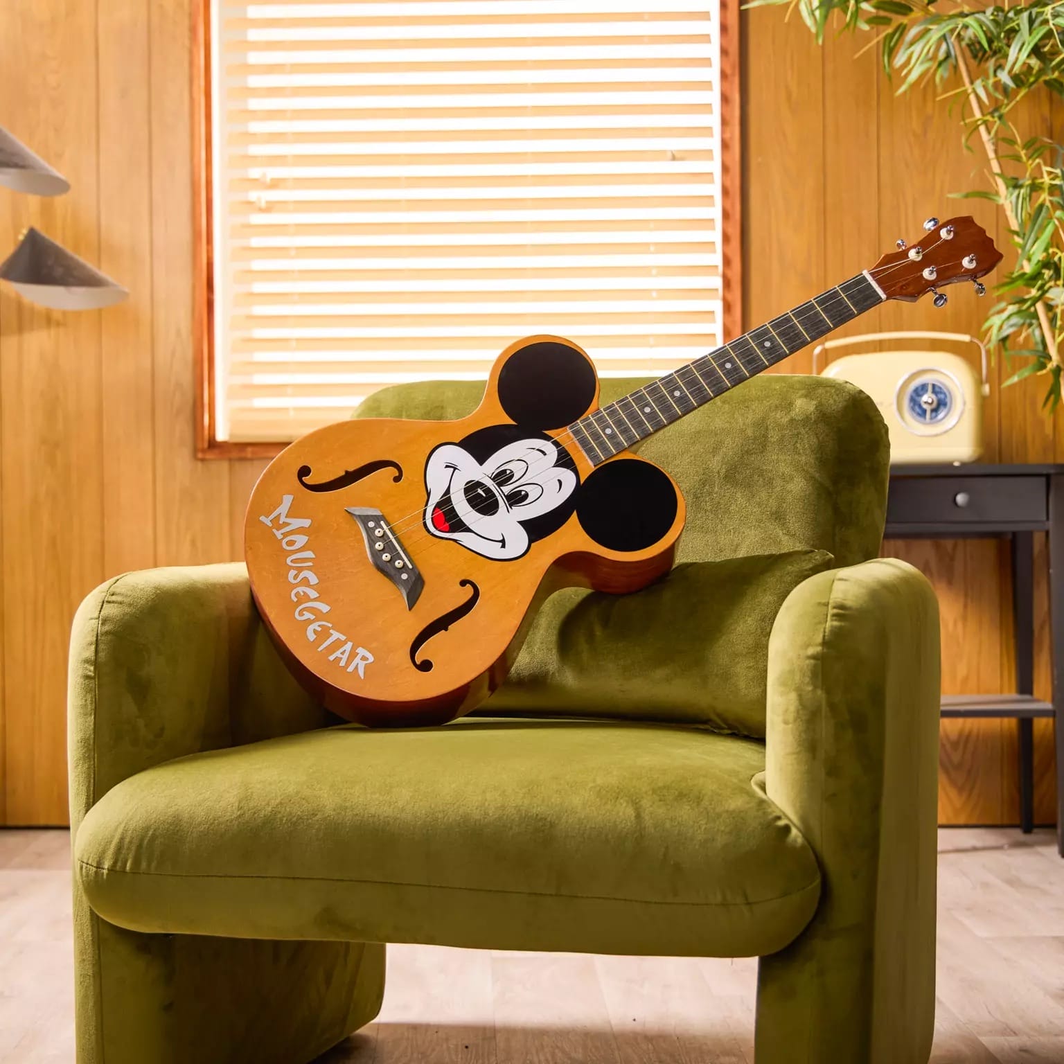 A Disney Store-Exclusive guitar with a Mickey Mouse face rests on a green armchair in a cozy, wood-paneled room.
