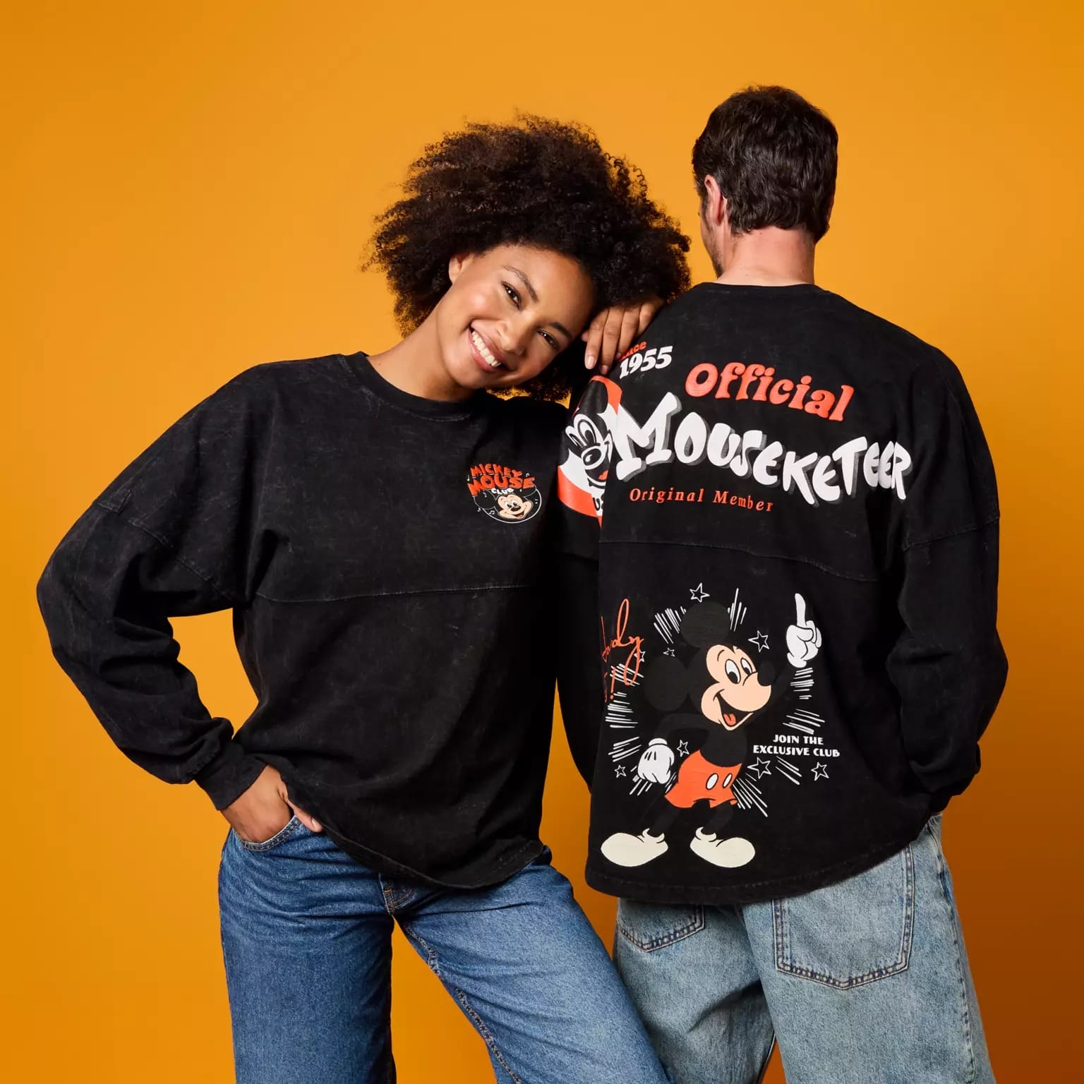 Two people in Disney Store-Exclusive Mickey Mouse Club Spirit Jerseys pose against an orange background, front and back.