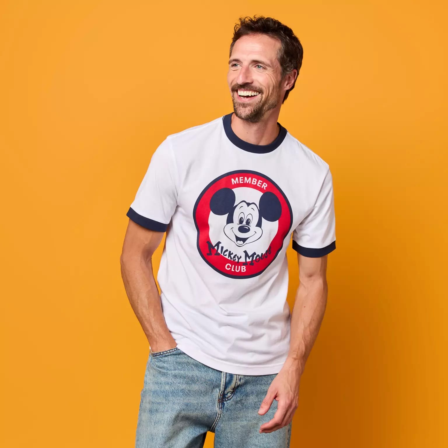 Smiling man in jeans and a Disney Store-Exclusive Mickey Mouse Club 70th Anniversary T-shirt stands on orange background.