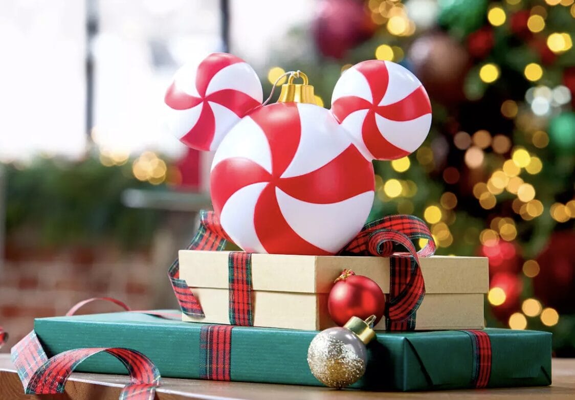Festive Disney ornaments and gifts surround a Peppermint Swirl Mickey Mouse before a glowing Christmas tree, evoking park magic.