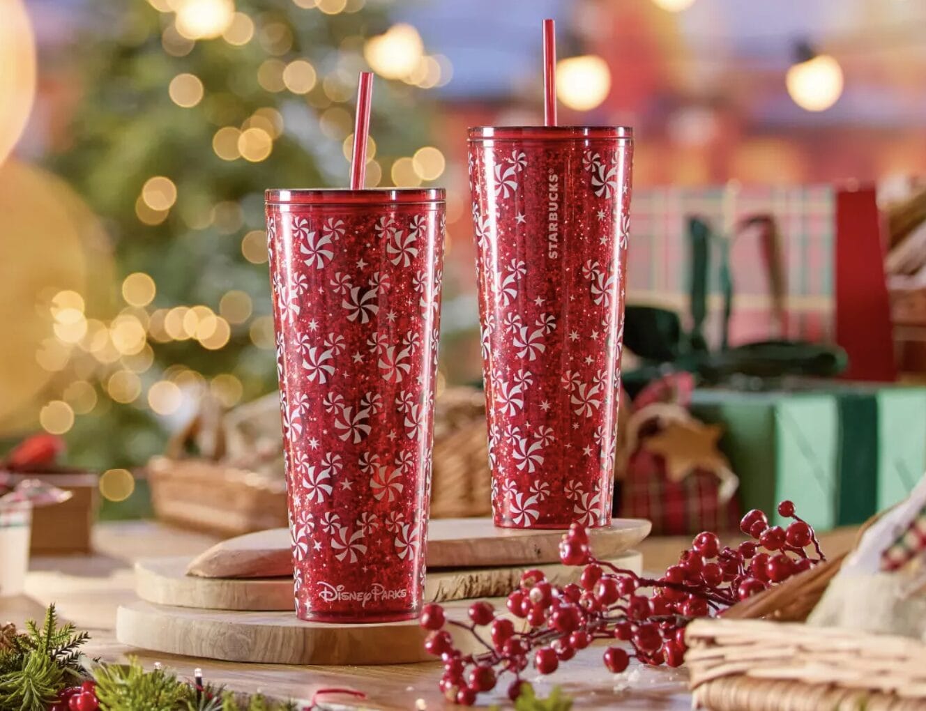 Two red Starbucks holiday tumblers with peppermint swirl and snowflake designs sit by a Christmas tree near Disney-themed presents.