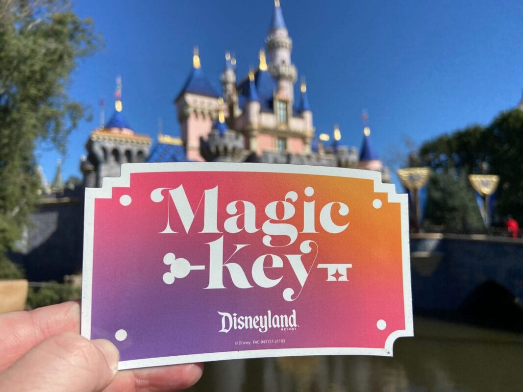 A hand holds a bright "Magic Key" magnet in front of Sleeping Beauty castle at Disneyland Park.
