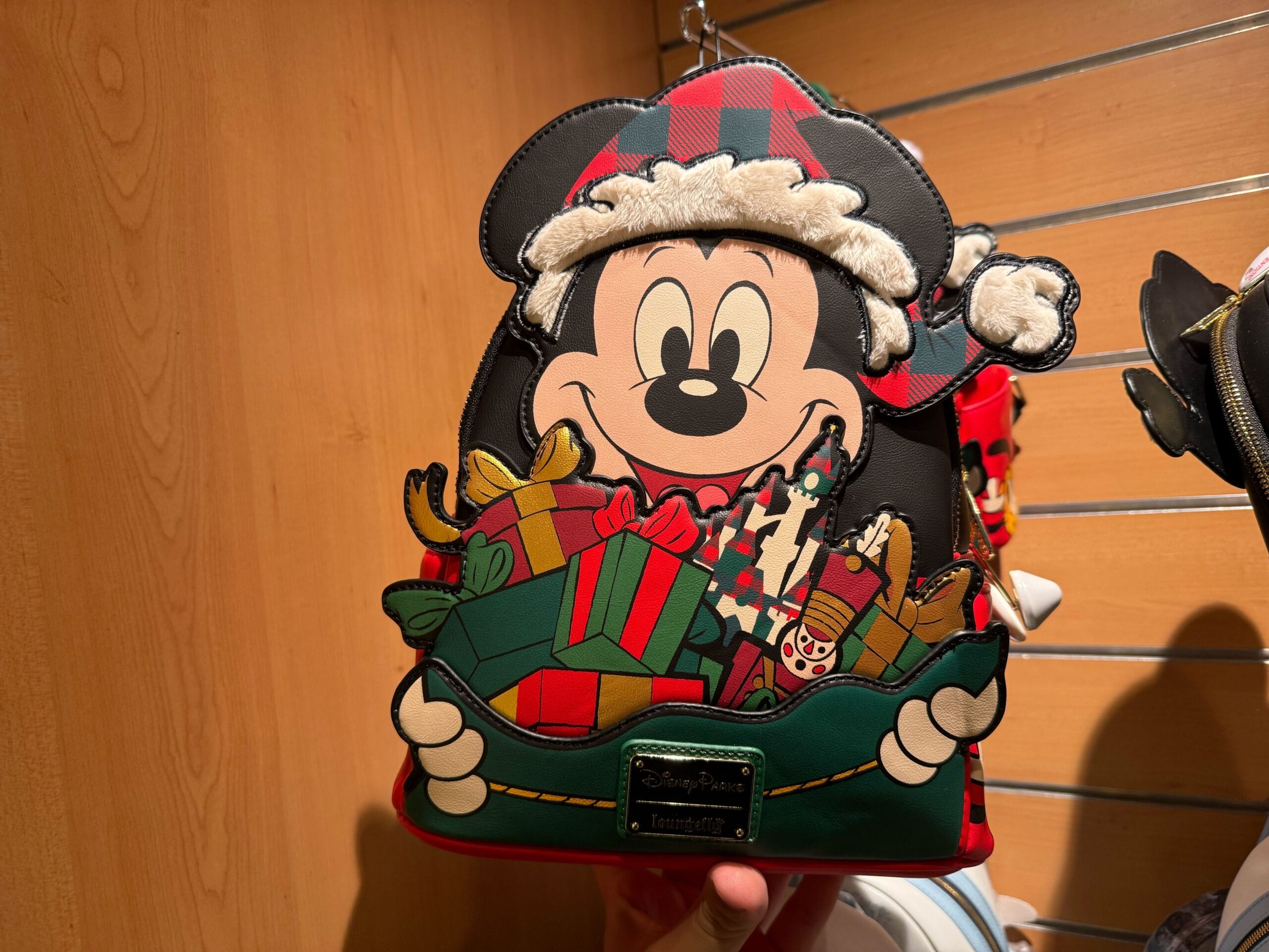 A festive Christmas Loungefly Backpack featuring Mickey Mouse in a Santa hat and holiday gifts, displayed on a wooden shelf.
