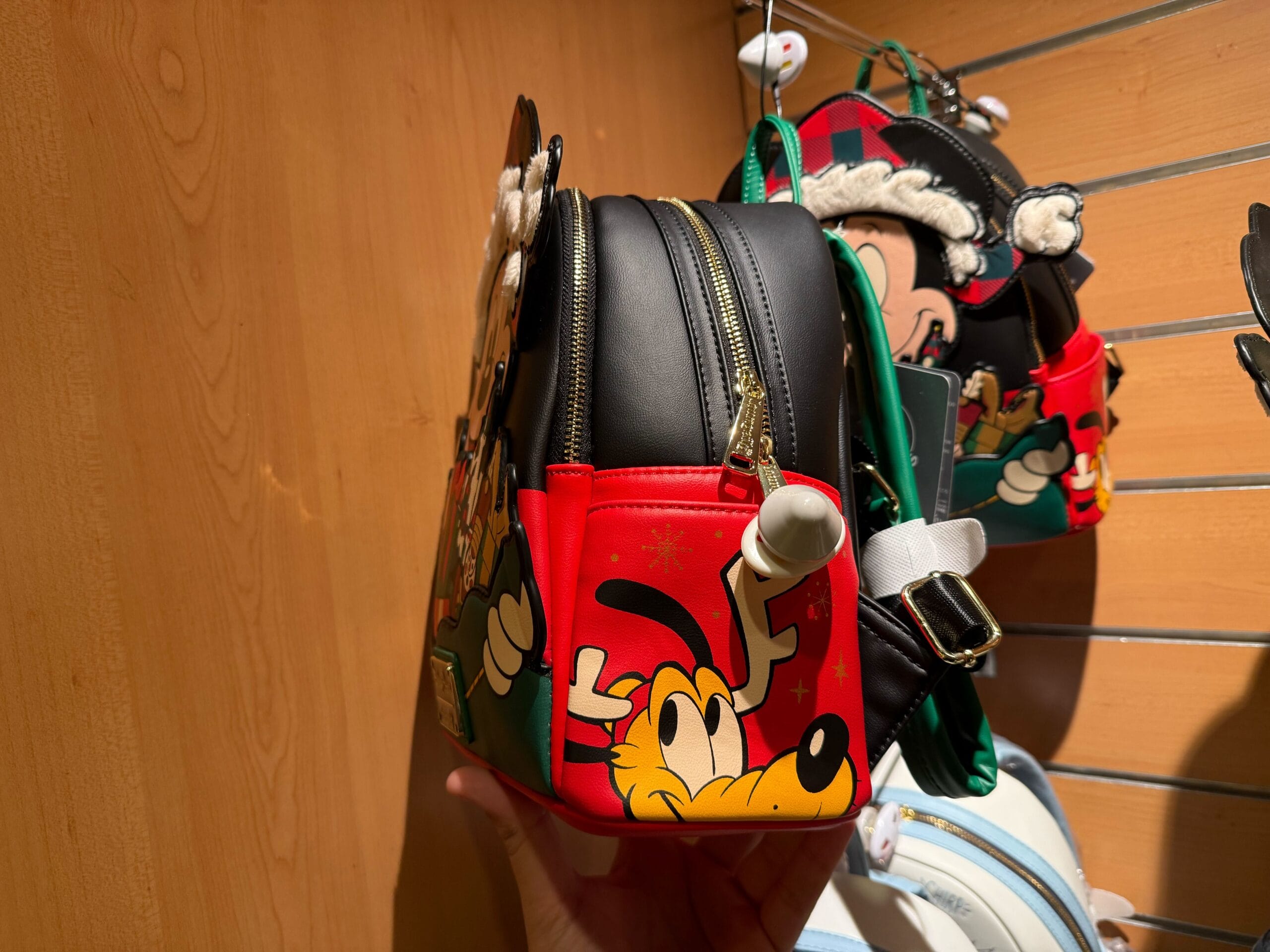 A hand holds a colorful Christmas Loungefly Backpack with a cartoon Pluto design, displayed among other bags in the store.