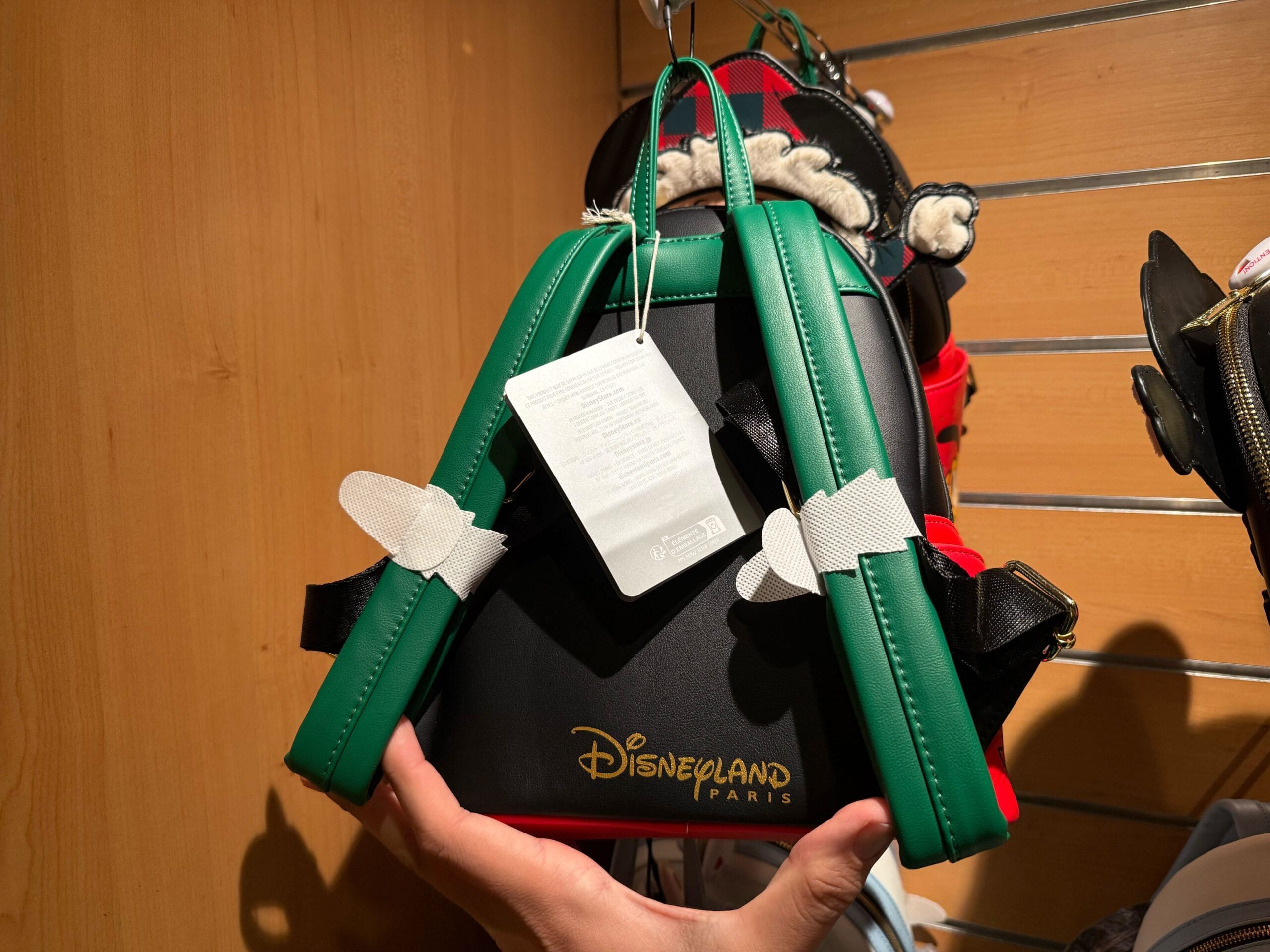 A hand holds a Disneyland Paris Christmas Loungefly Backpack with green straps and white tag, displayed on a wooden shelf.