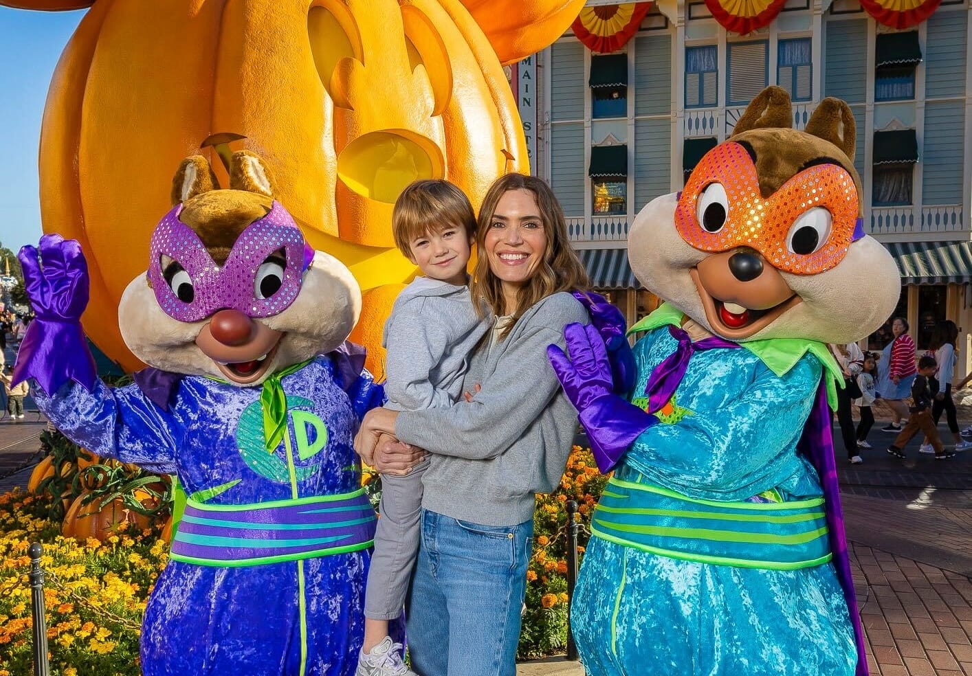 A woman with a child poses with two Disney chipmunks in festive outfits by a large pumpkin at a Disney park, no castle visible.