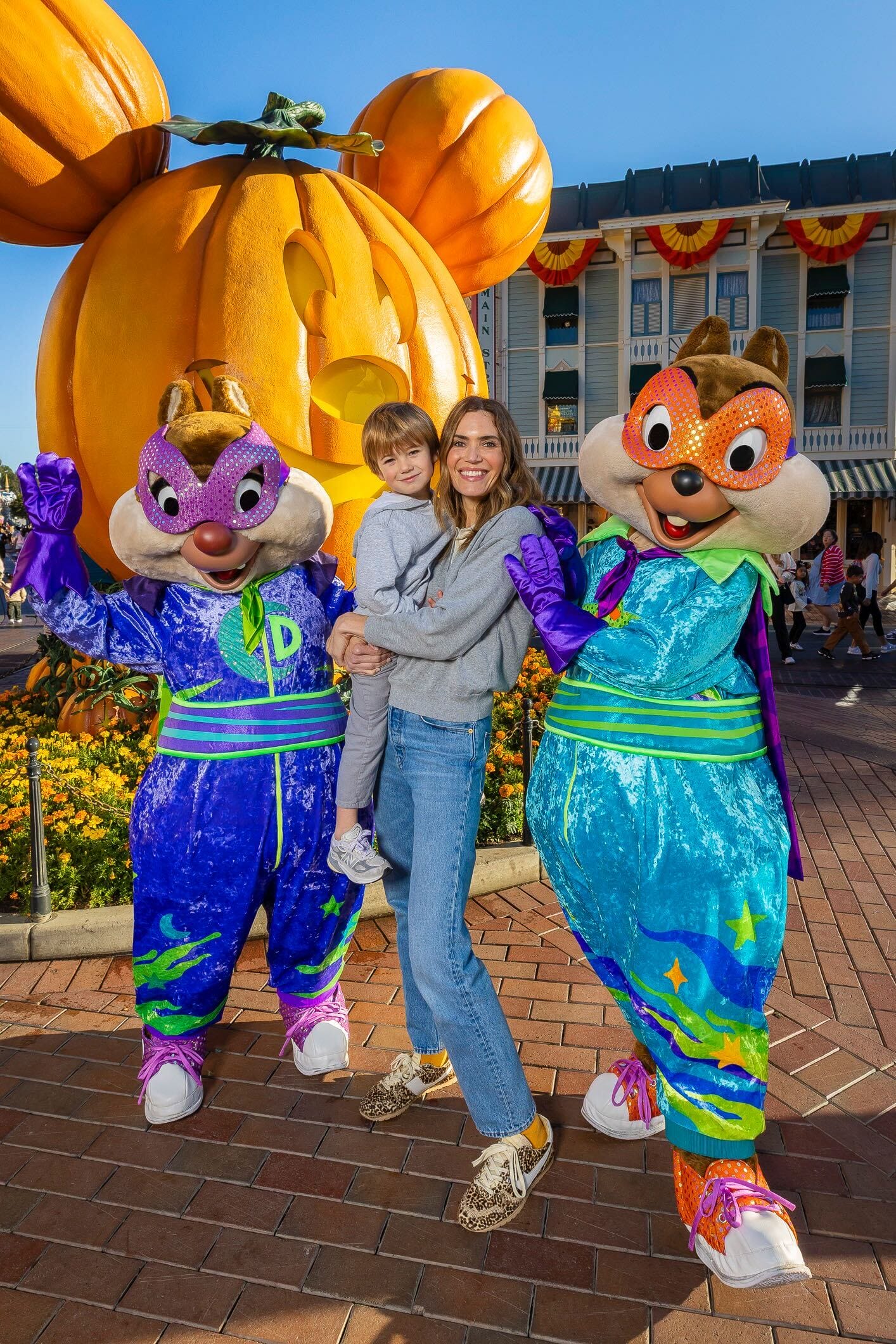 A woman with a child stands with Chip 'n Dale mascots in front of a giant pumpkin at a Disney park decorated for Halloween.