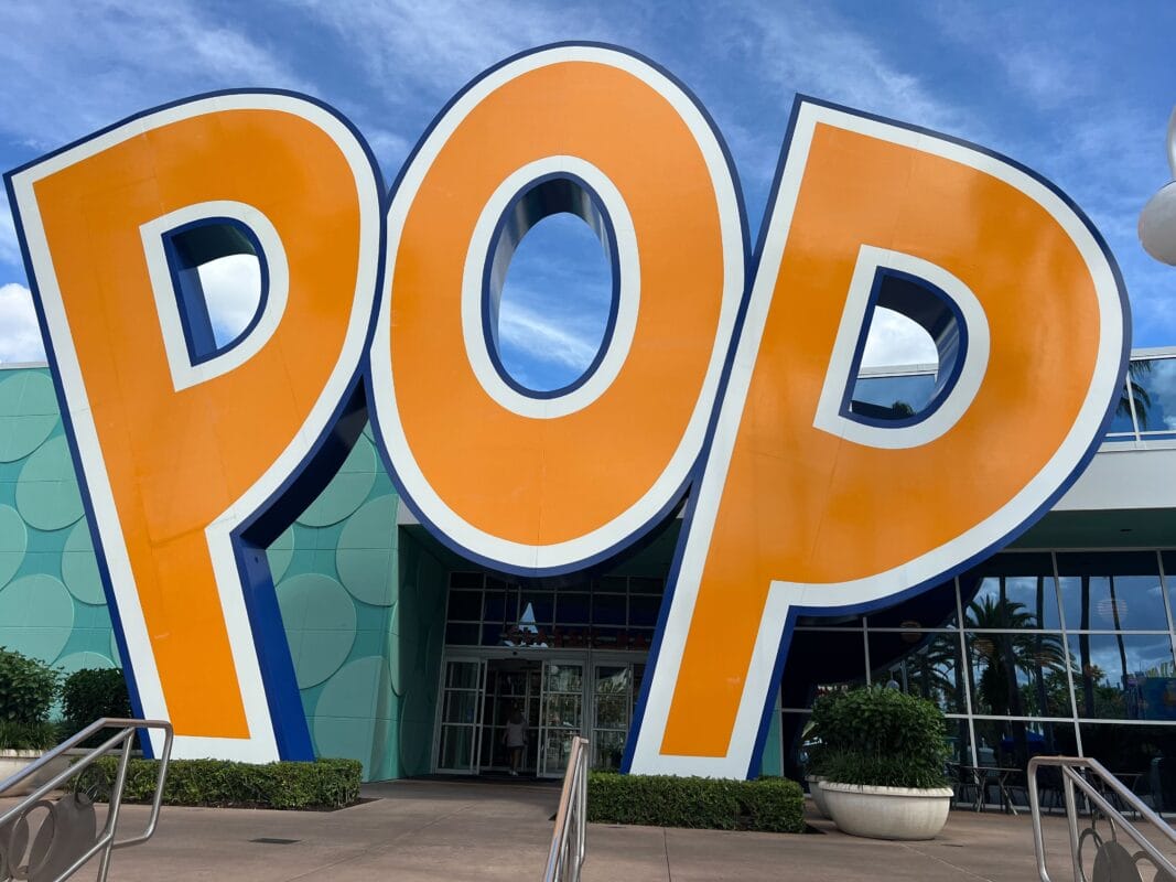 Large orange "POP" letters in front of Disney’s Pop Century Resort, teal walls behind, blue sky—no castle unlike any Disney park.