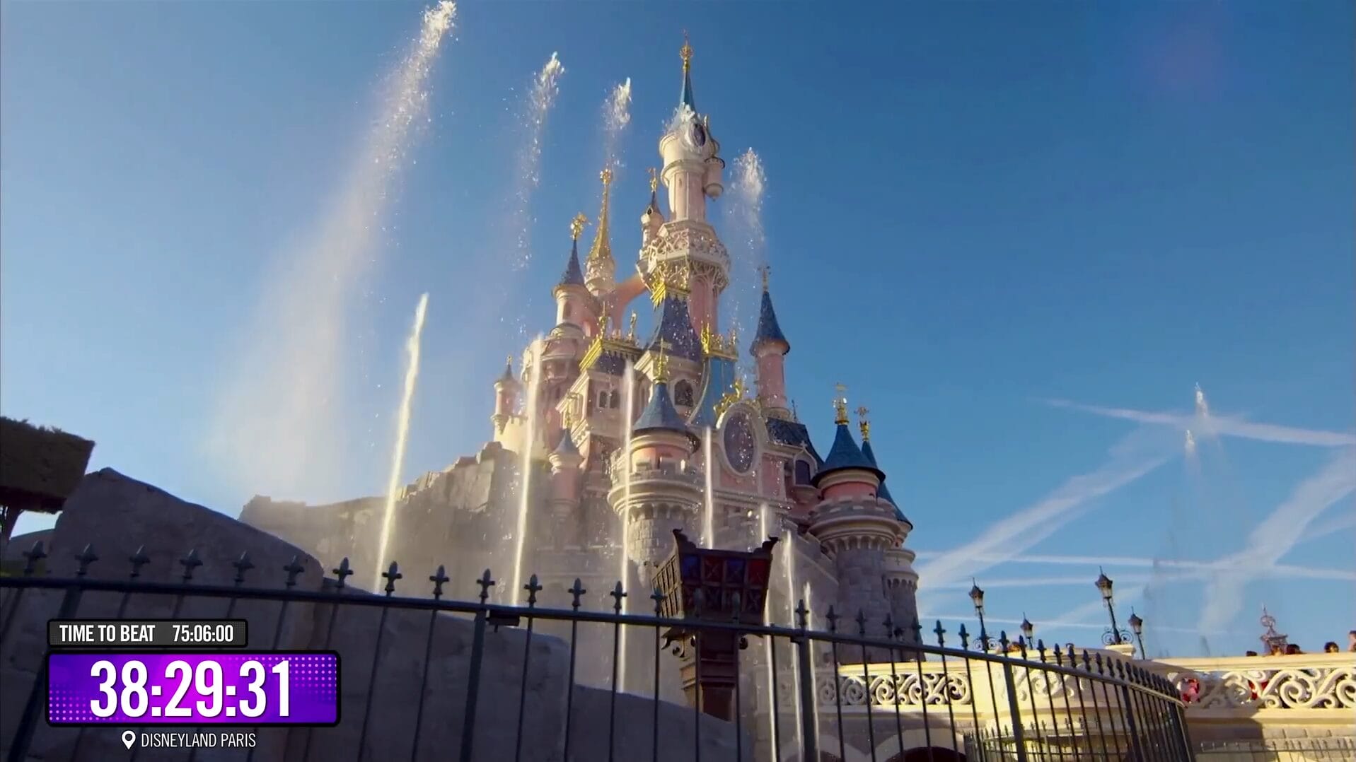 Disney Park castle with fountains, seen through a gate under blue skies; 75 Hours timer by Dude Perfect at bottom left.